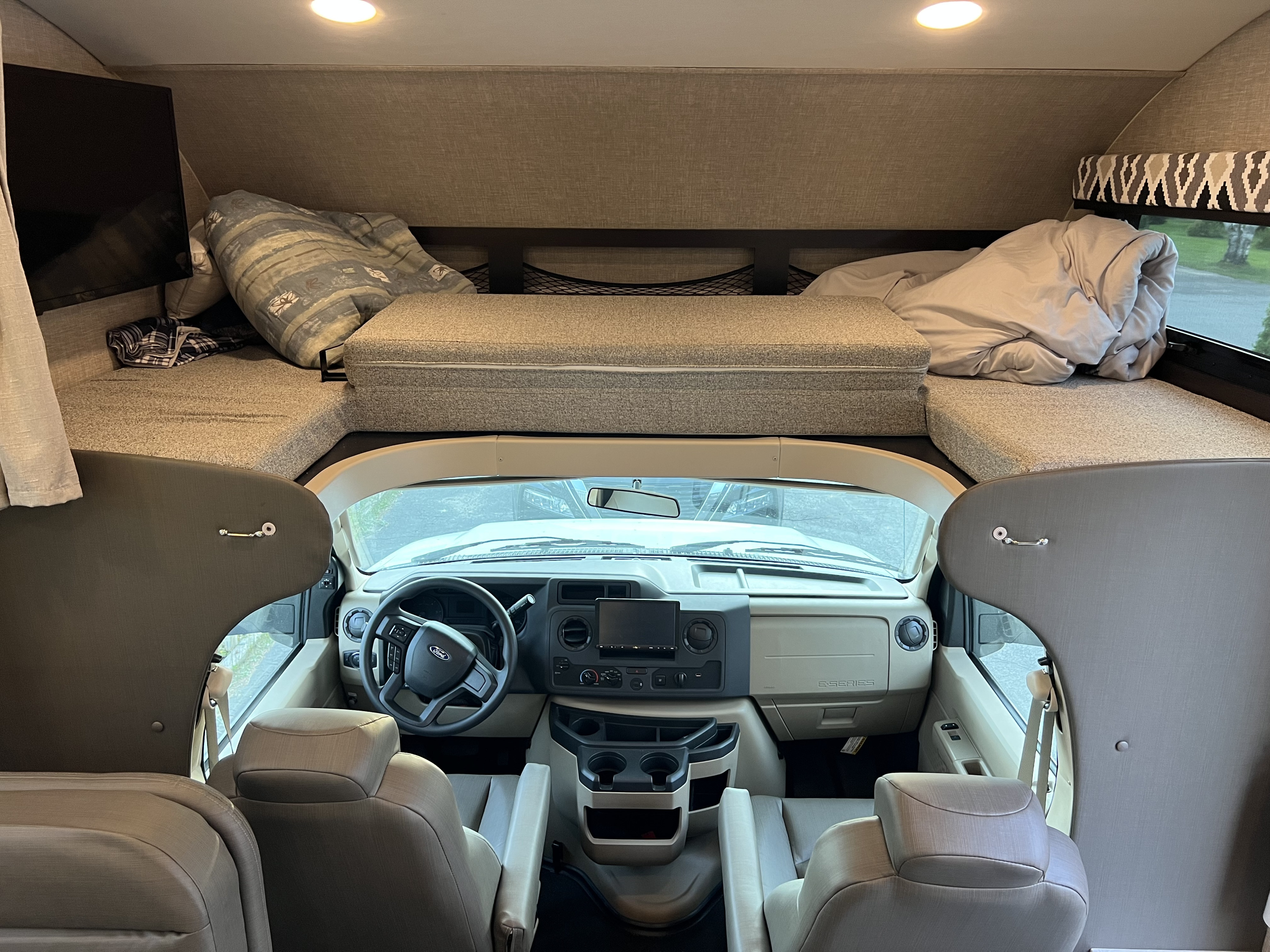 Inside a comfy RV rental: view of the cab-over bunk with bedding & TV, looking down to the Ford cockpit and seating area.. Jayco Greyhawk 2022