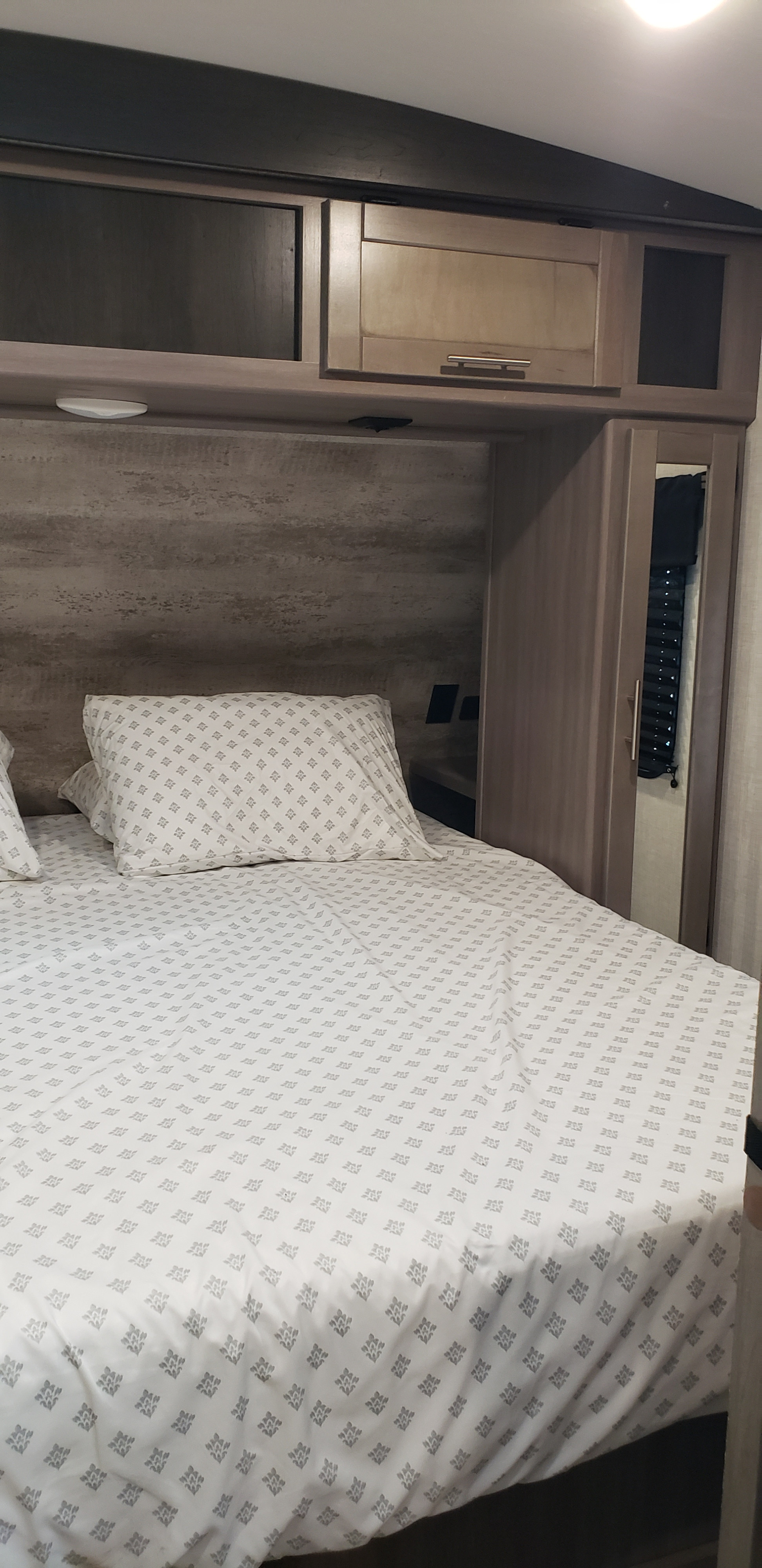 View of an RV rental bedroom showing a bed with patterned sheets, wood-look headboard, and overhead cabinets.. Keystone Sunset Trail 2022