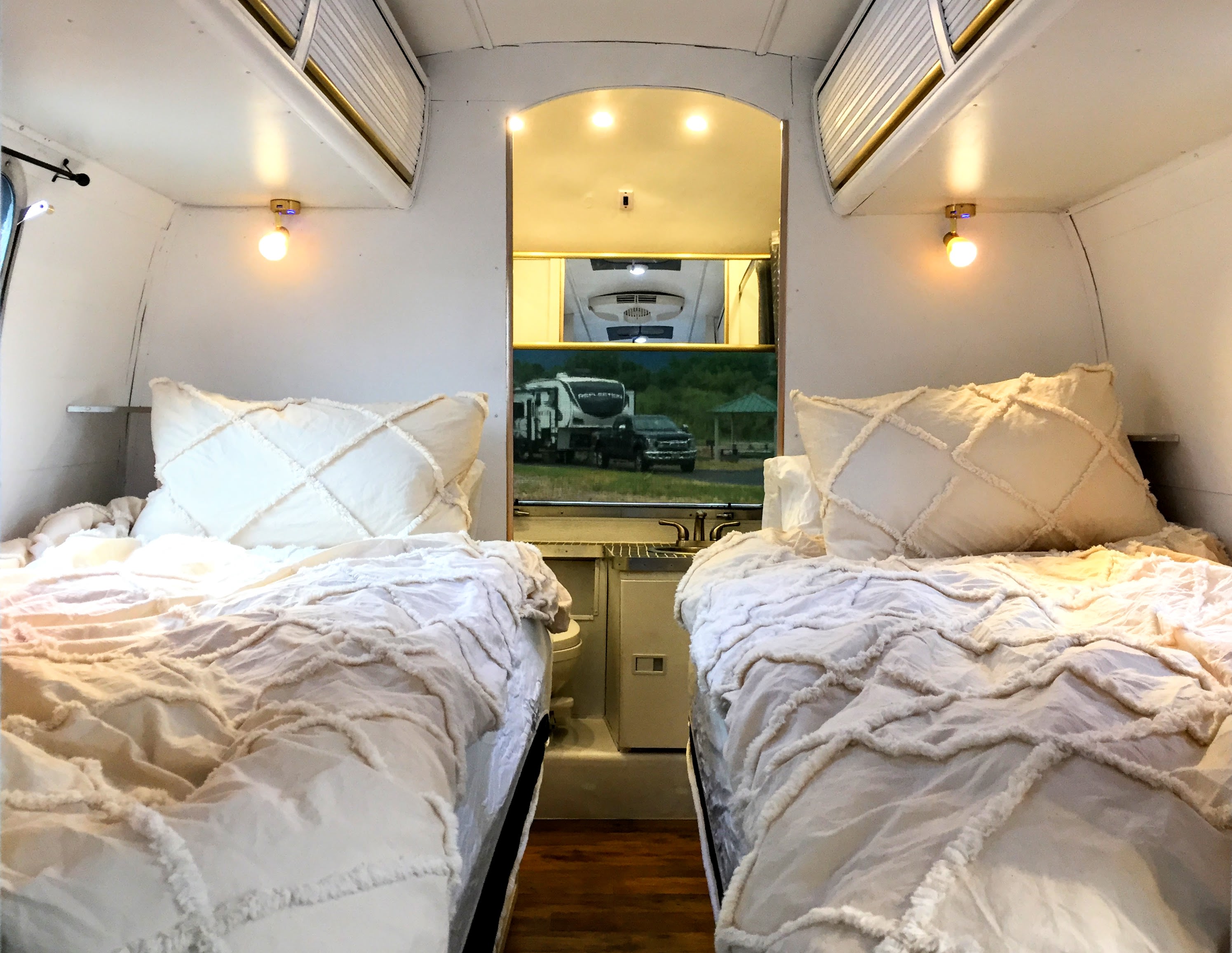Relaxing RV rental interior with two twin beds featuring textured cream bedding. Includes a view of the bathroom/kitchenette area.. Airstream Trade Wind 1976