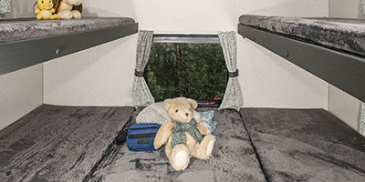 Inside an RV rental, two sets of comfy bunk beds with grey bedding & playful stuffed animals. Window views trees outside.. Venture Stratus 2022
