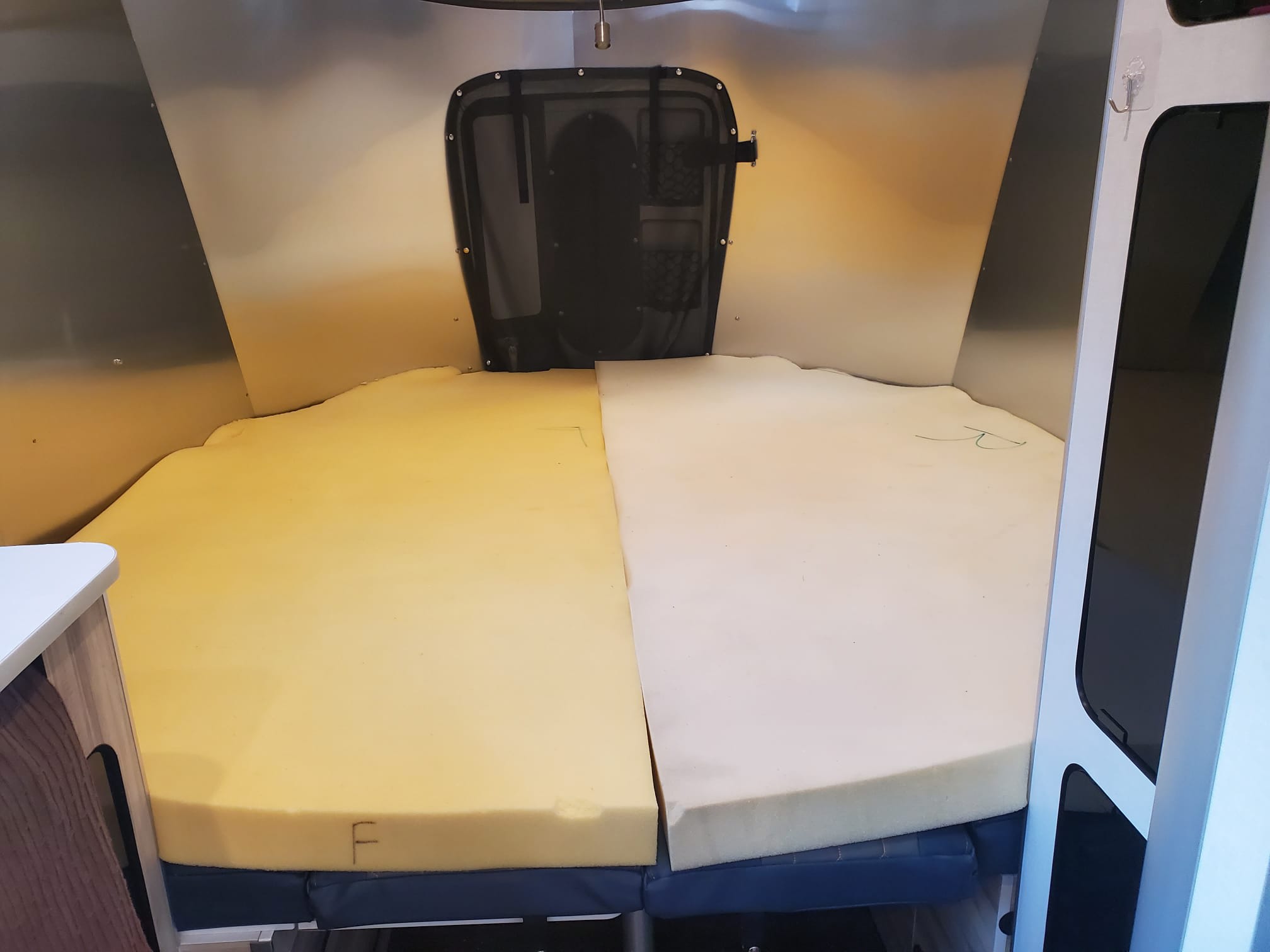 RV rental interior featuring a two-tone foam bed/mattress setup. Metallic walls and a mesh rear vent define this compact camper space.. Airstream Basecamp 2022