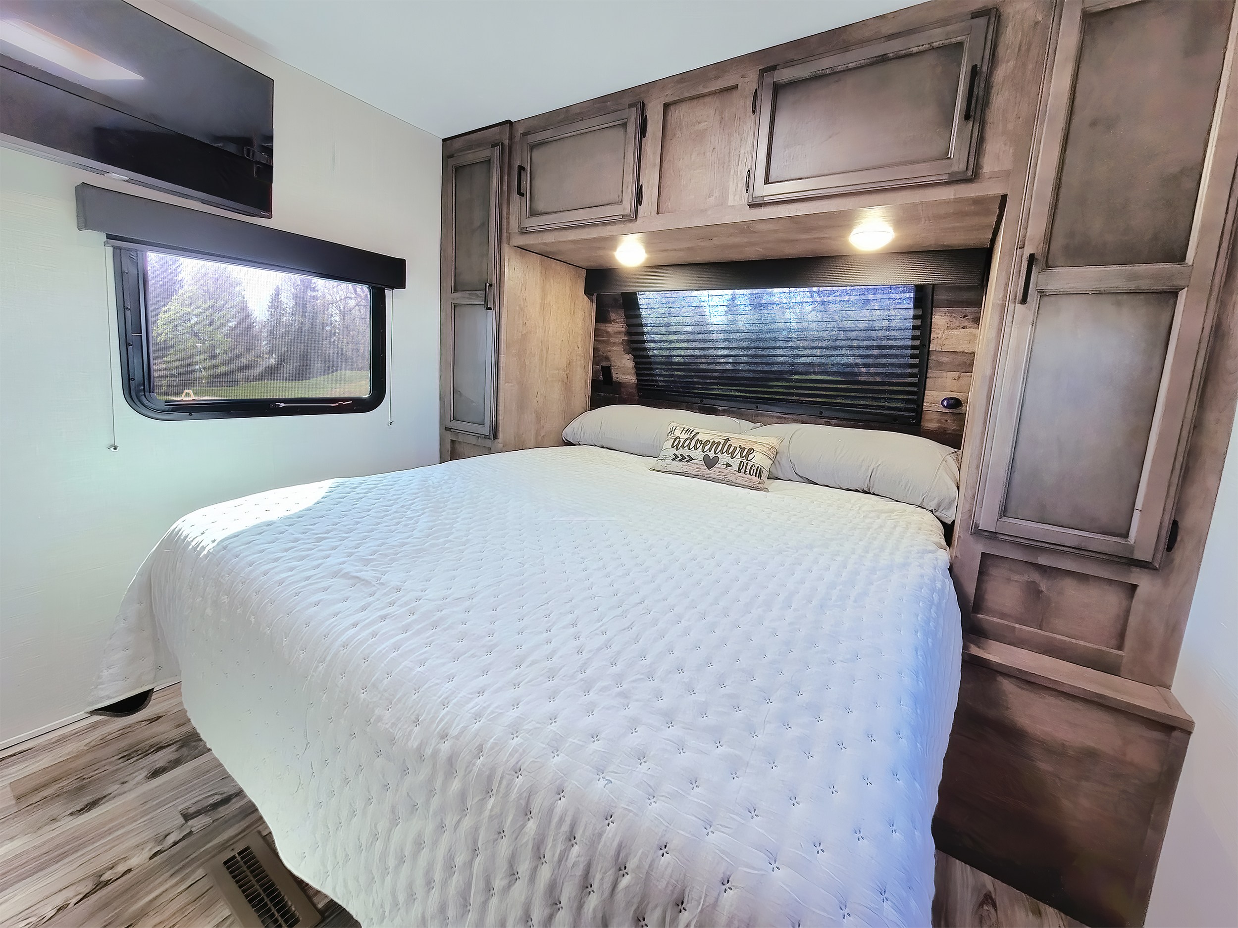 Cozy RV bedroom for your next adventure. Features a large bed, rustic wood cabinets, window view. Ideal for an RV rental experience.. Venture Rv Sporttrek 2022