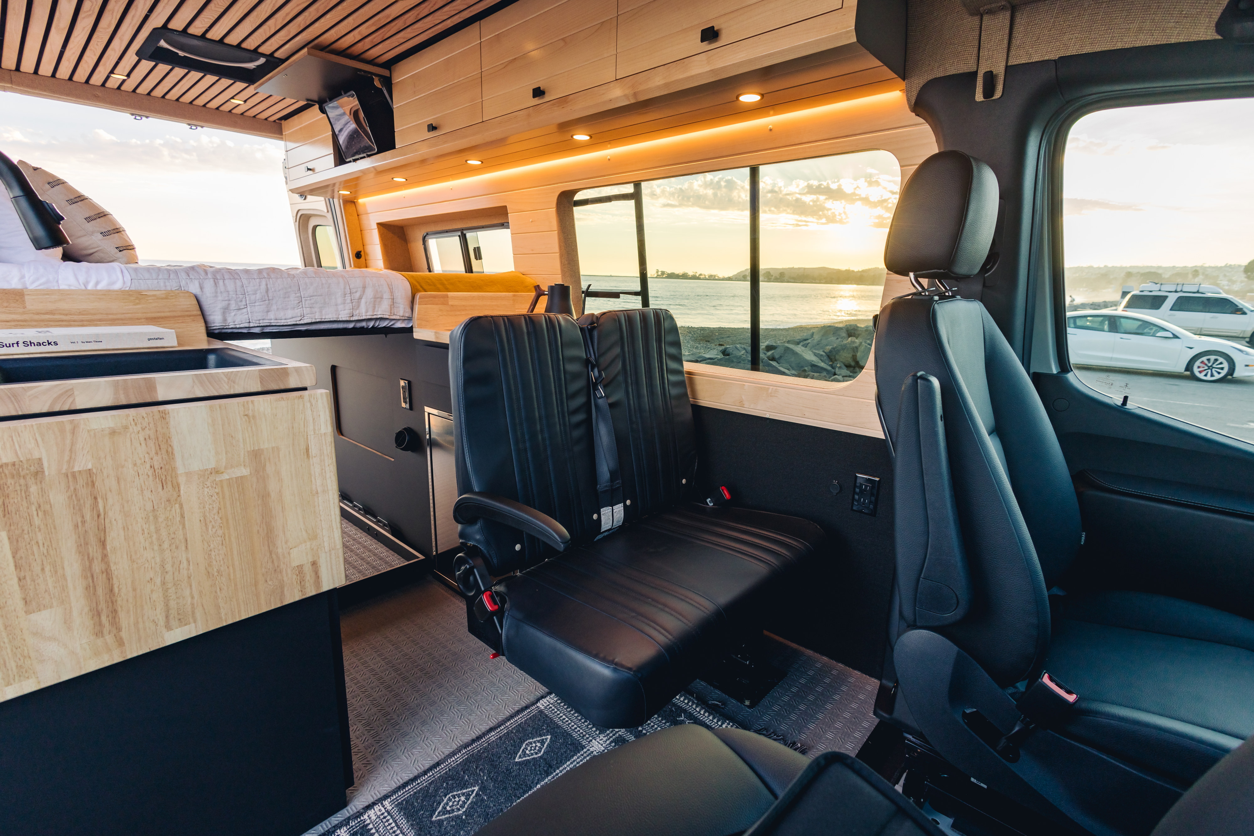 Spacious, wood-paneled RV rental interior with bed, seating, and stunning sunset ocean views from your camper van.. Mercedes-Benz Sprinter 2023