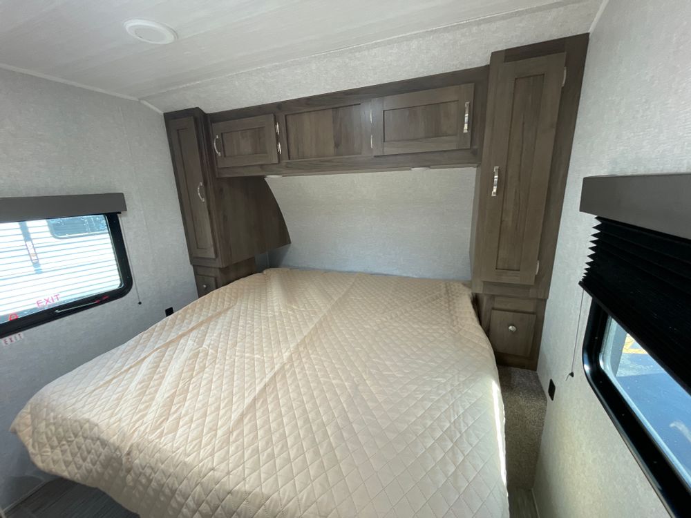 Compact RV rental bedroom featuring a large bed with a light-colored quilted comforter, overhead, and side cabinets.. East to West 25KRB 2022