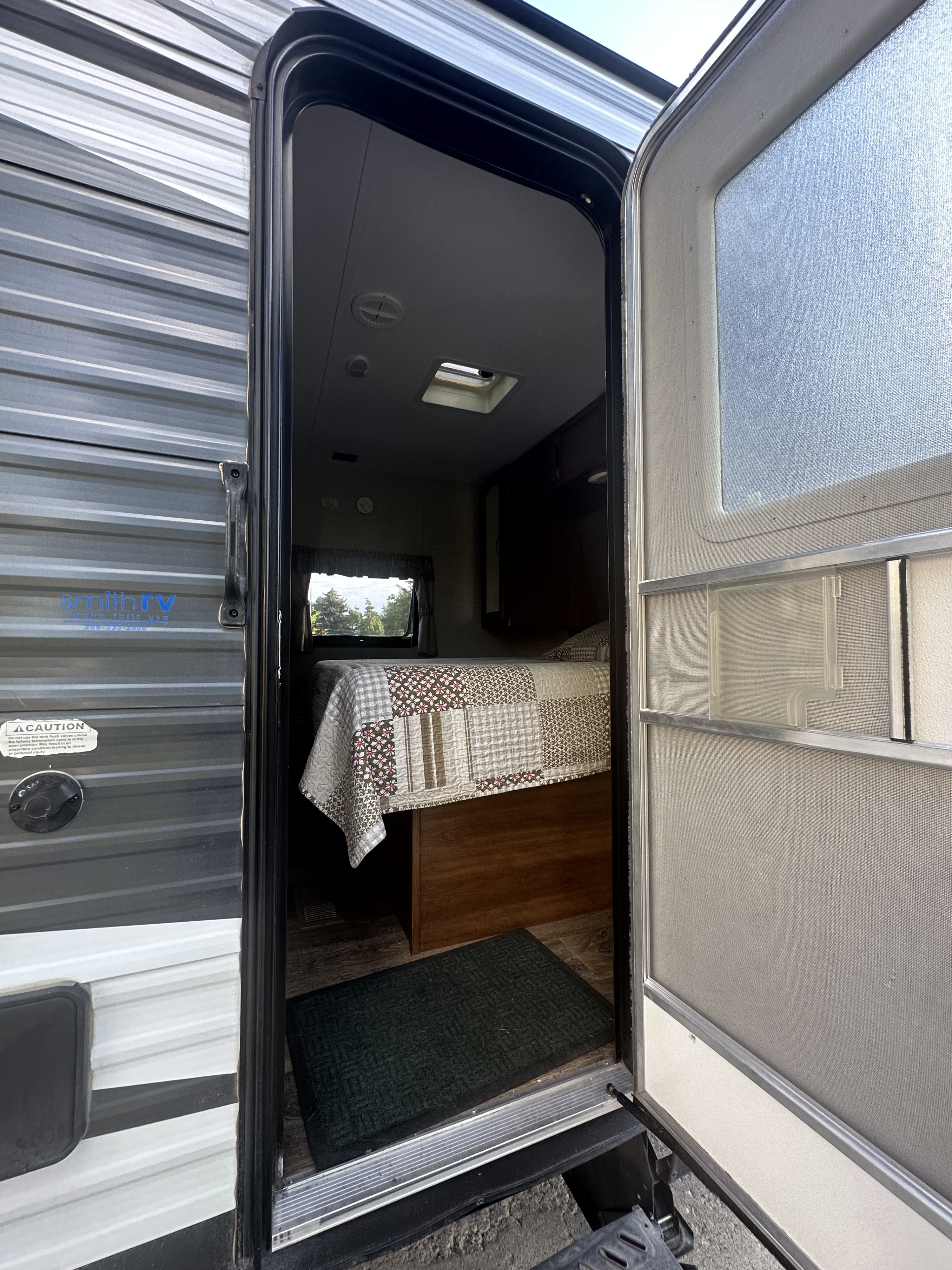 Open door of an RV rental revealing its cozy bedroom with a patterned quilt, window view, and floor mat.. Keystone RV Springdale 2016