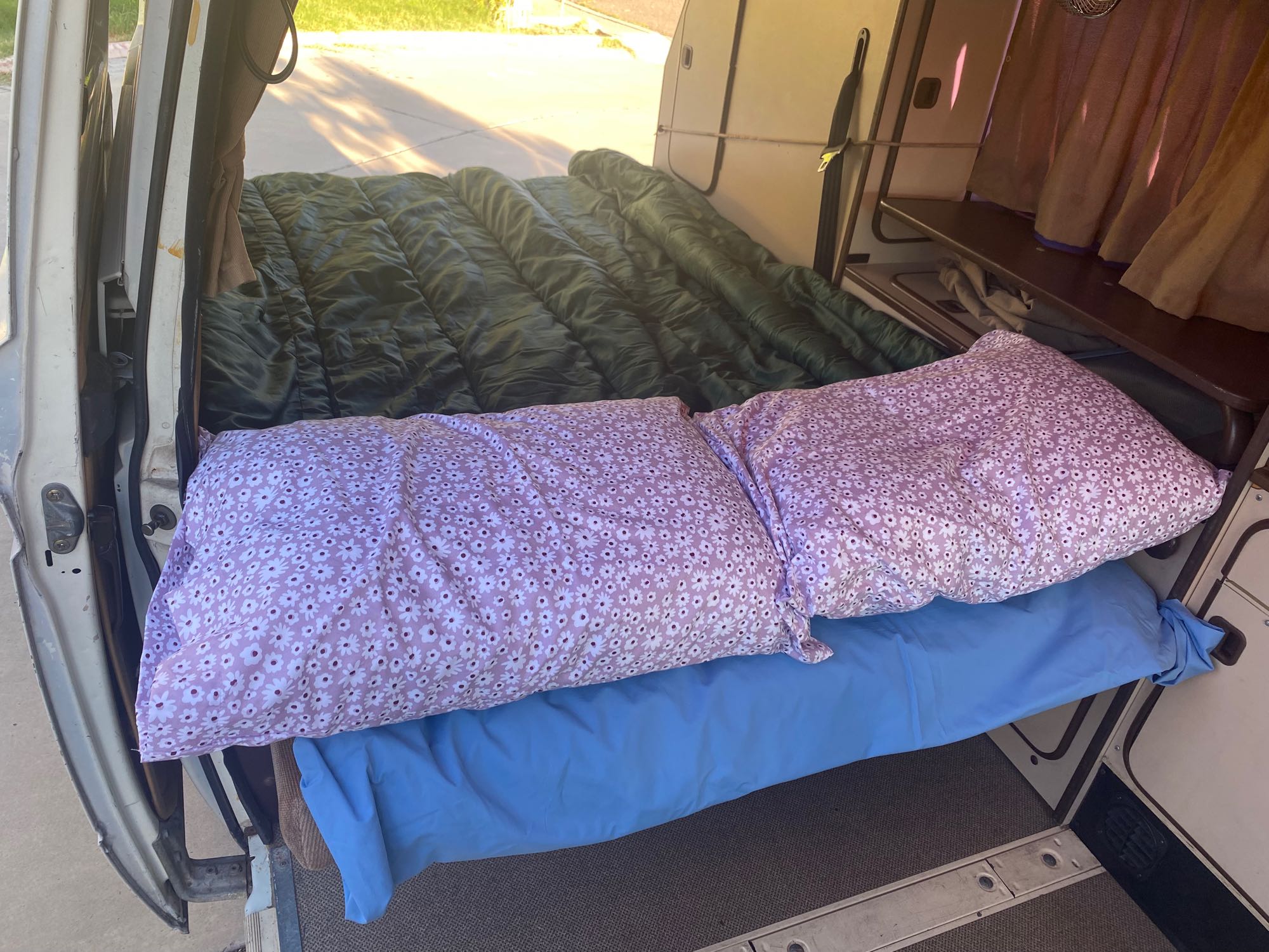 Experience comfort in an RV rental. This camper van features a green comforter, blue sheets, and purple floral pillows.. Volkswagen Westfalia 1985