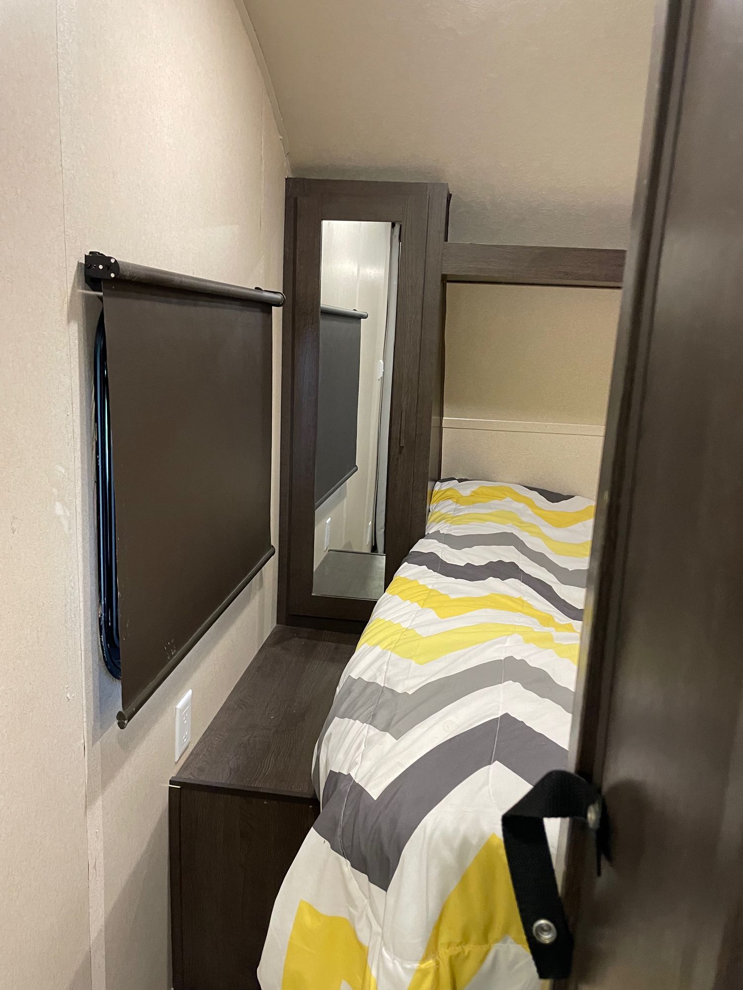 This RV rental bedroom offers a cozy bed with a yellow and gray striped comforter, dark window blind, and built-in storage.. Forest River Wildwood X-Lite 2019