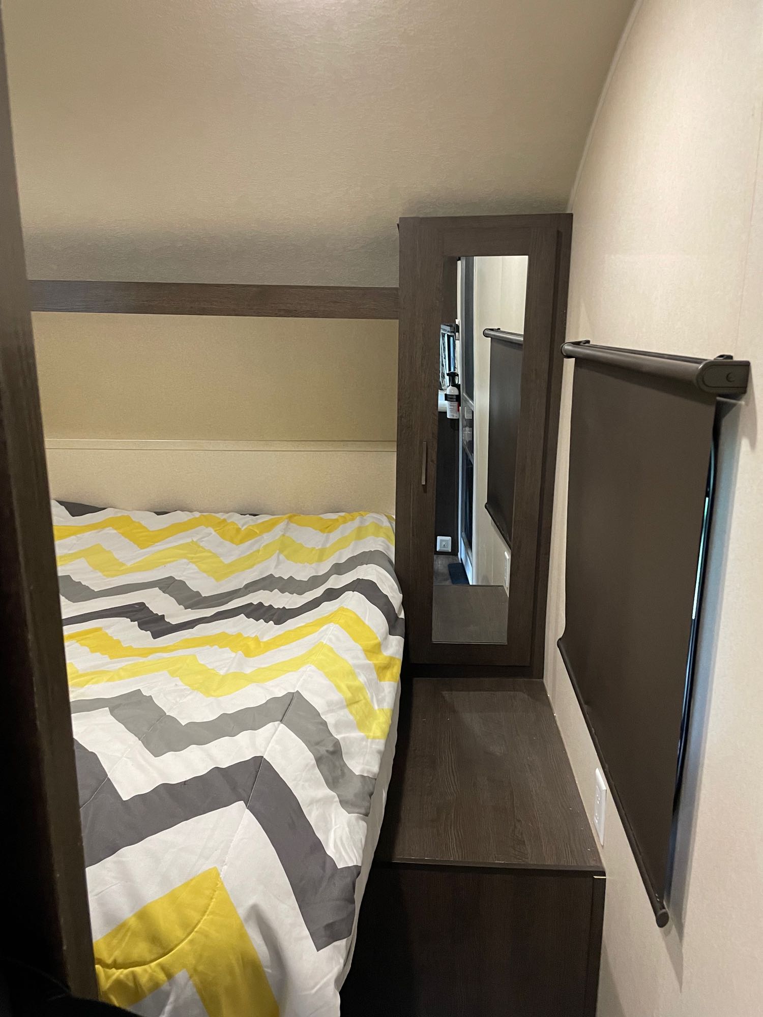 Inside an RV rental: a compact bedroom with a yellow/grey chevron bedspread, mirrored wardrobe, and roller blind.. Forest River Wildwood X-Lite 2019