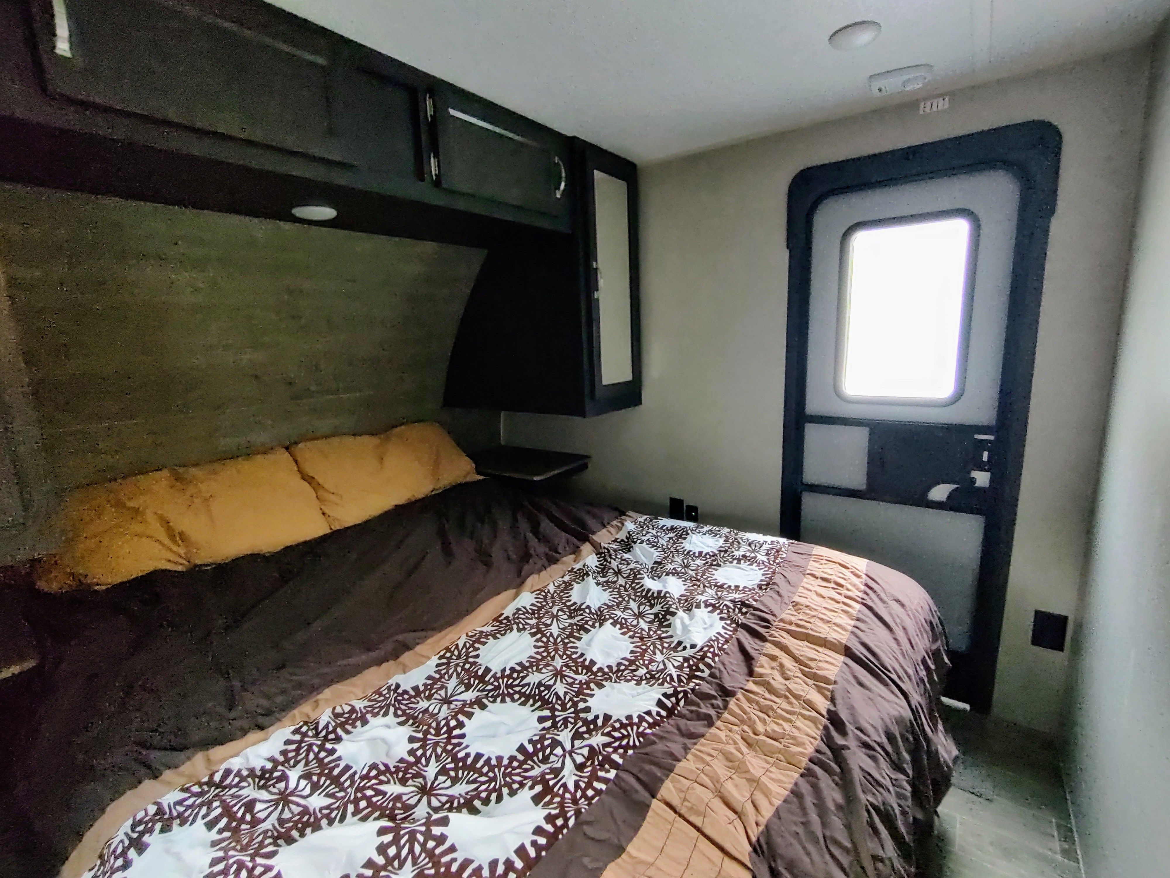 RV rental bedroom interior featuring a bed with patterned brown & white bedding, overhead cabinets, and an exit door.. Palomino Puma 2020