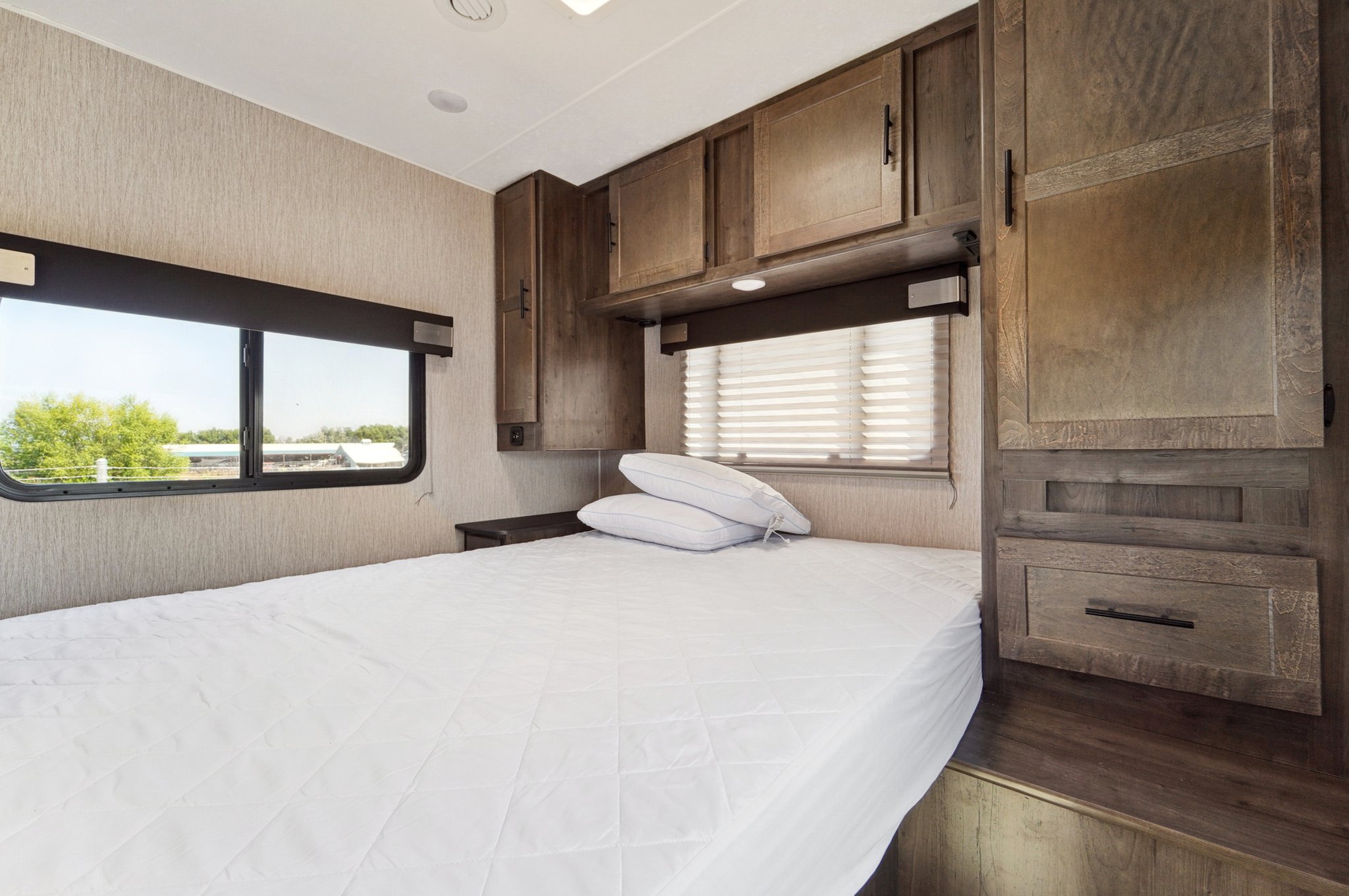 Experience comfort in this RV rental bedroom: white bed, dual windows, and ample wood storage. Ideal for road trip relaxation.. Coachmen Leprechaun 2021