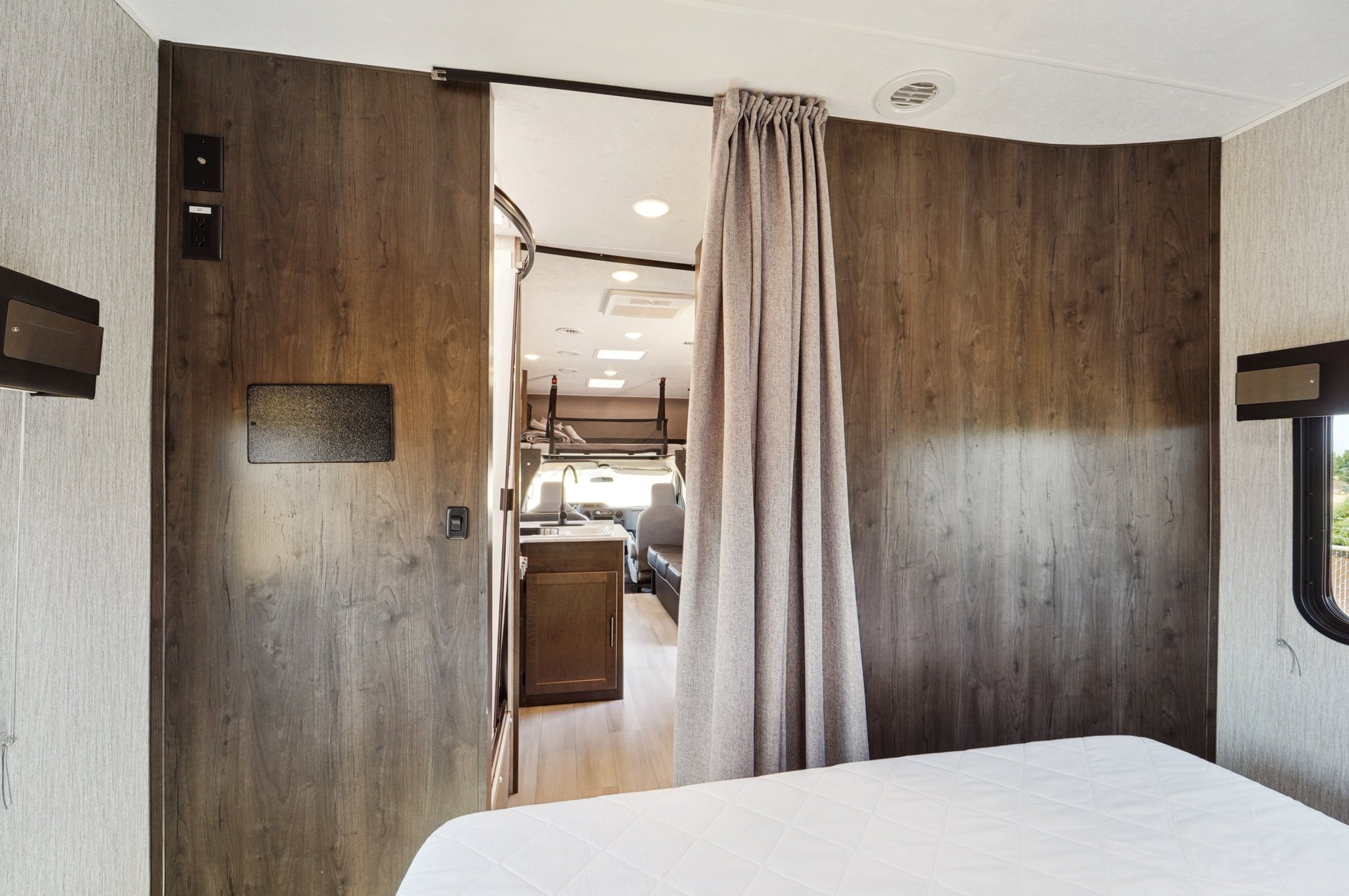 View from an RV rental bedroom showing white bed, wood paneling, and a privacy curtain opening to the living area.. Coachmen Leprechaun 2021