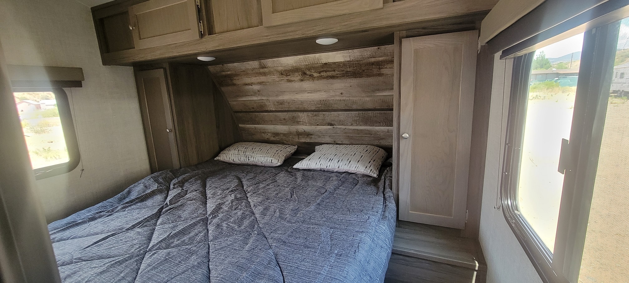 Inside an RV rental: a cozy bedroom with a patterned comforter, wood headboard, cabinets, and bright windows.. Gulf Stream Gulf Breeze 2023