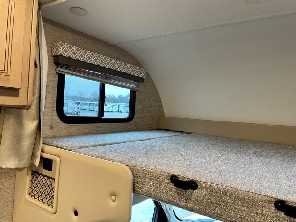 Inside an RV rental: a cozy over-cab bunk bed with a window and storage, ready for your next adventure.. Thor Coleman 2022