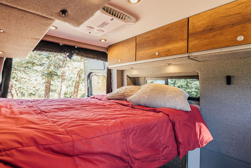 Spacious RV rental interior featuring a comfy red bed, wood cabinets, AC unit, and forest views through windows.. Storyteller Overland Storyteller Overland Class B 2022