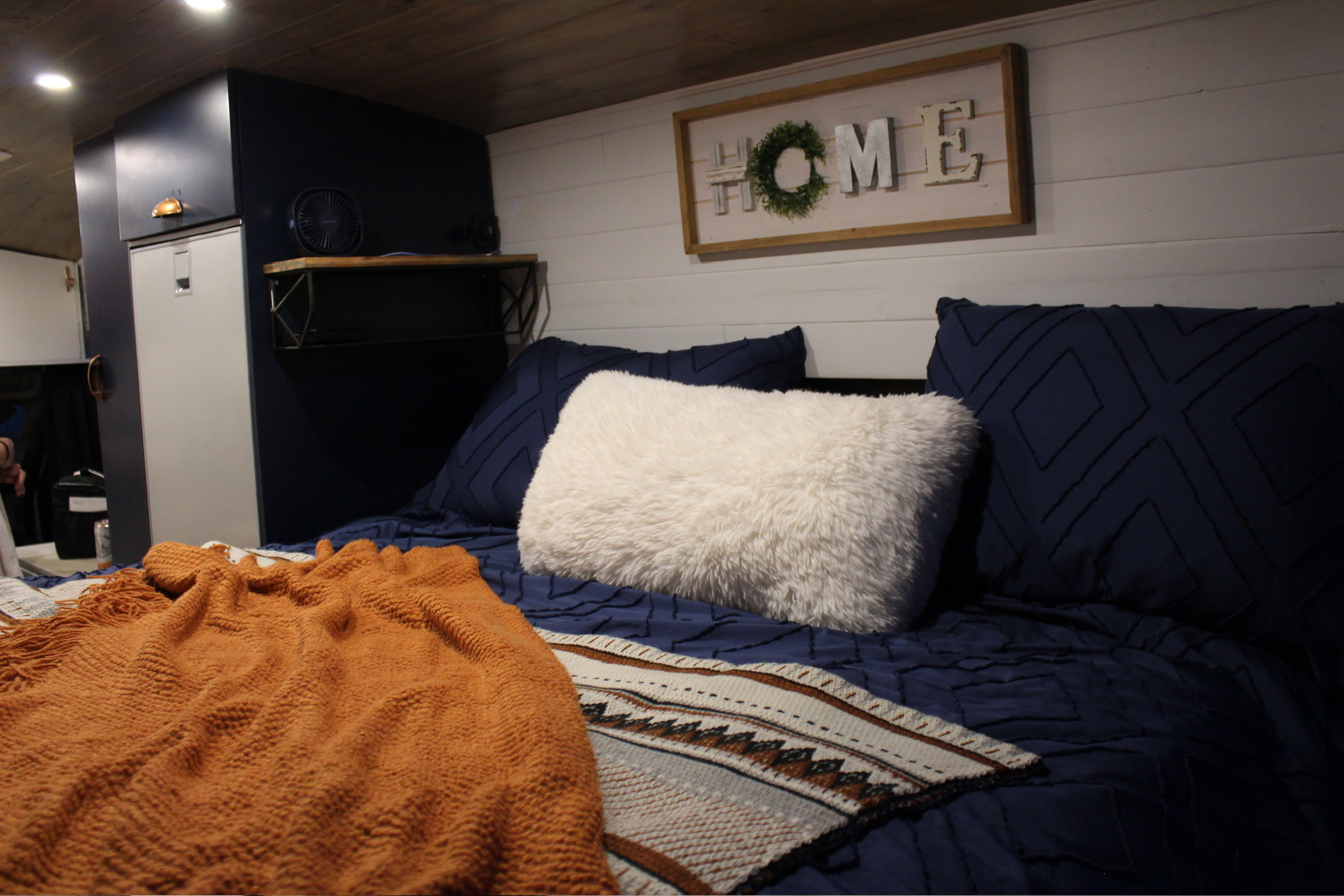 Cozy RV rental bedroom with navy blue bedding, a fluffy white pillow, orange blanket, and a HOME sign.. Ford Transit 2019