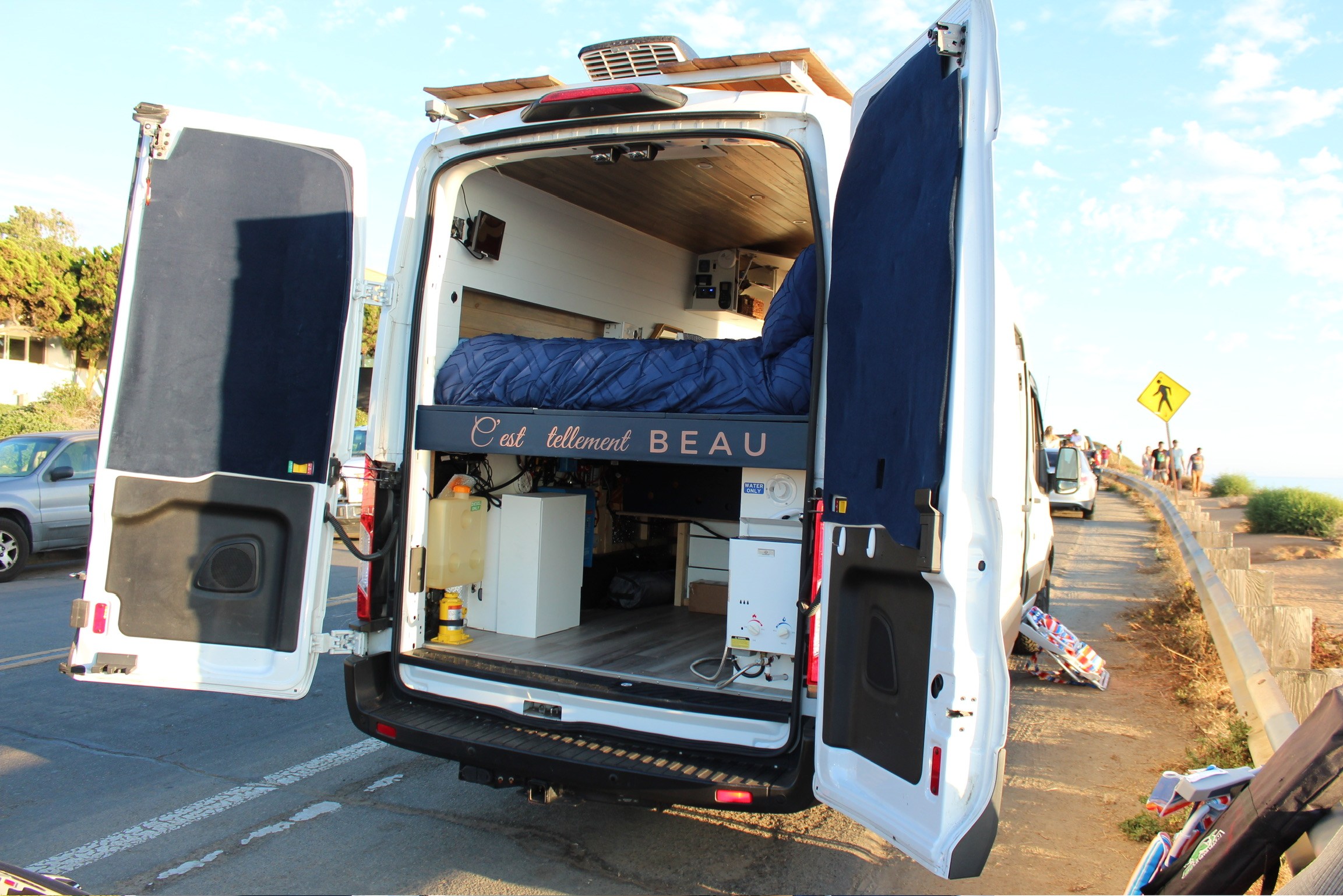 Open rear doors of a customized white camper van, showcasing its cozy bed, wood interior, and utilities, perfect for an rv rental.. Ford Transit 2019