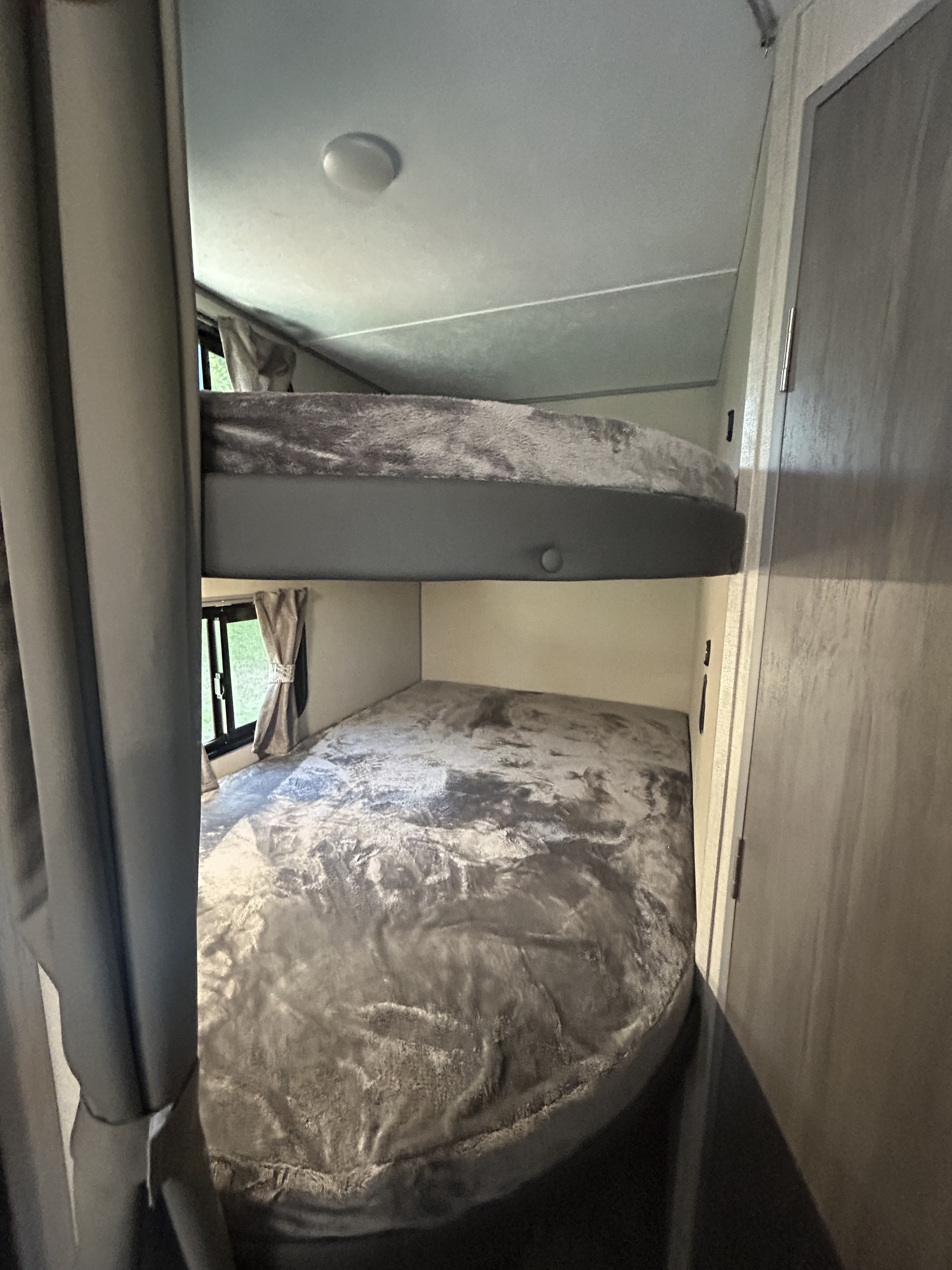 Inside an RV rental, showing cozy bunk beds with soft grey blankets. A compact sleeping area with windows and a side door.. Coachmen 184BH 2023