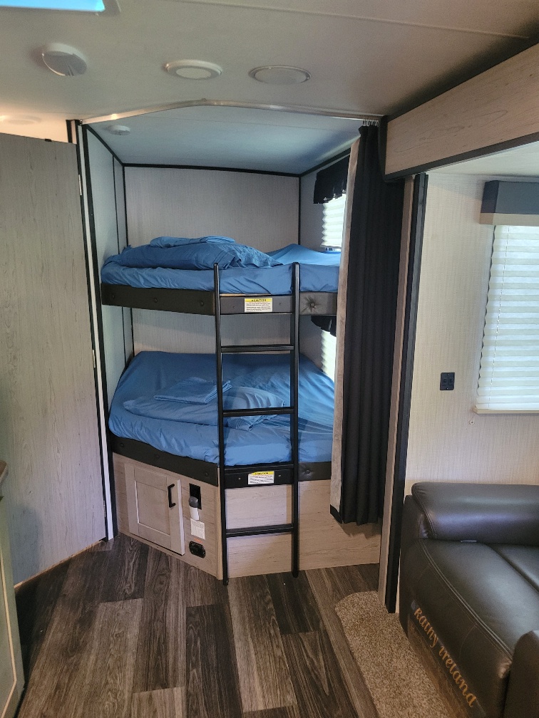 Inside an RV rental: compact bunk beds with blue sheets, a ladder, wood-look floor, and dark sofa.. Heartland RVs Mallard 2021