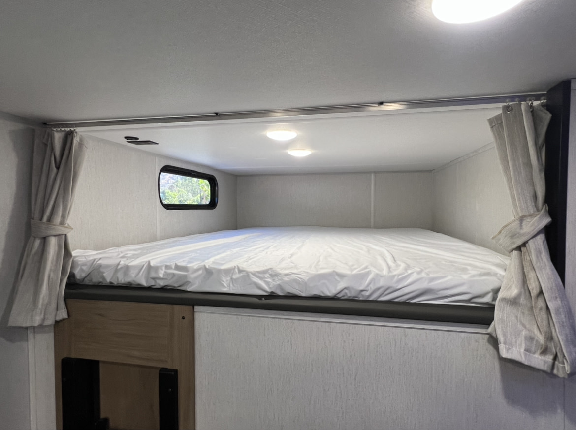 RV rental interior showing a comfortable bunk bed with a window, ceiling lights, and neat tied-back curtains.. Forest River Cherokee 2024