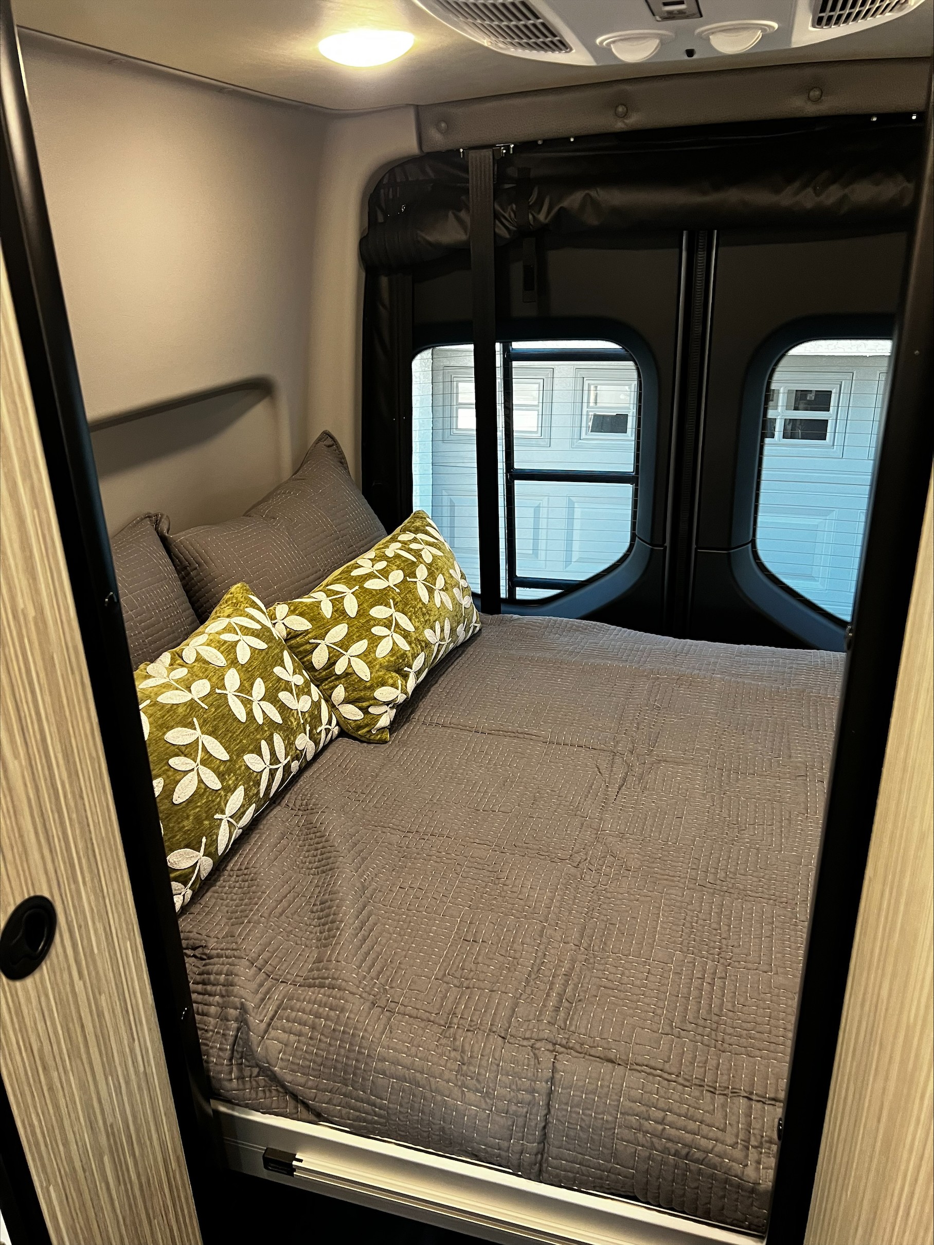 View into a compact RV rental sleeping area featuring a bed with grey bedding and two green leafy pillows.. Mercedes Winnebago Revel 2024