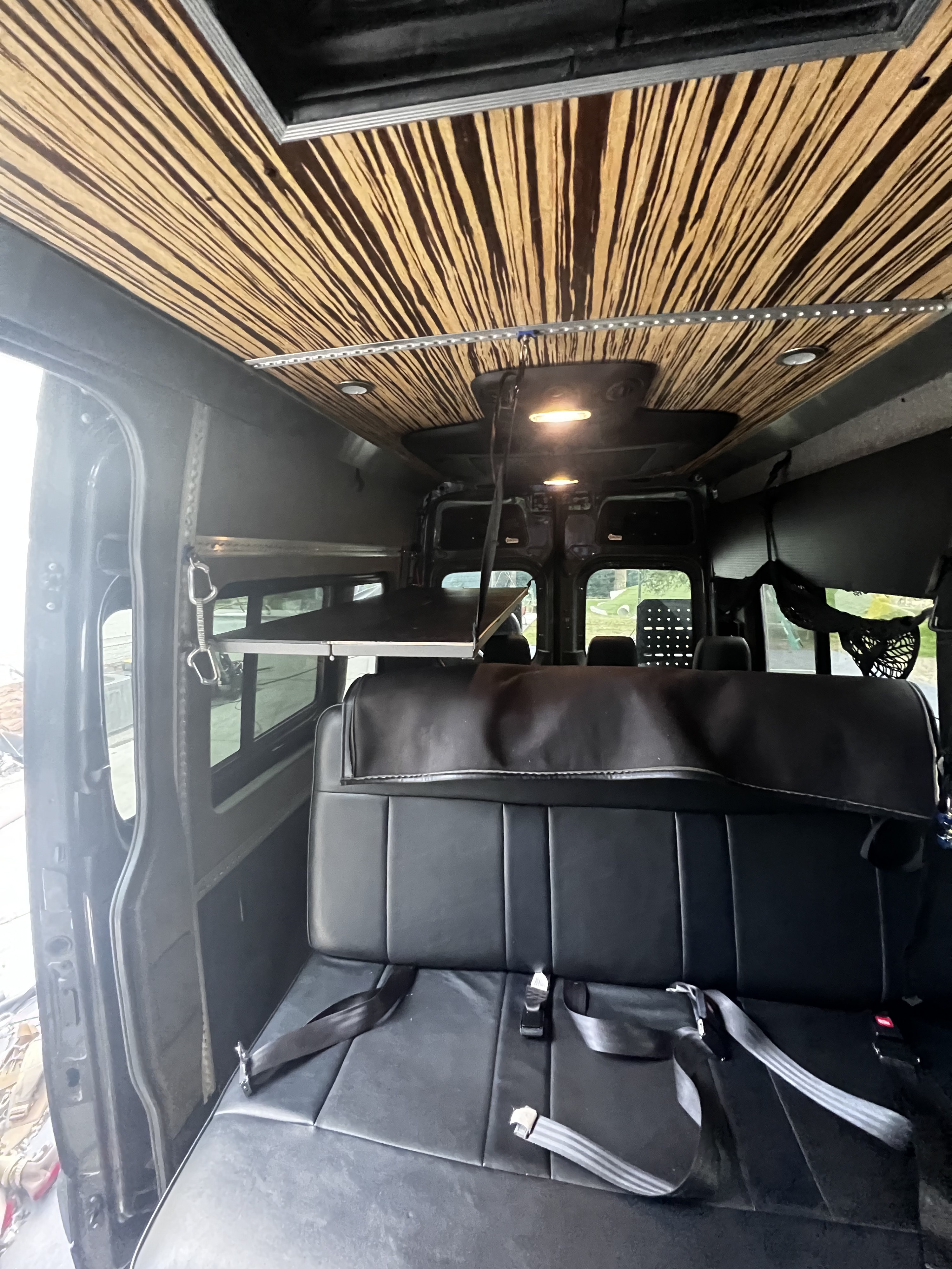 Inside an RV rental: a black bench seat and striped wood ceiling with LED lights. A fold-out bunk/shelf is on the left.. Mercedes-Benz Sprinter 2019