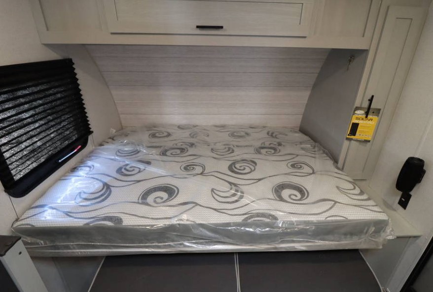 RV rental bedroom with a compact bed, new mattress in plastic, overhead cabinets, and a window blind for your trip.. Forest River Wildwood FSX Plantium 2022