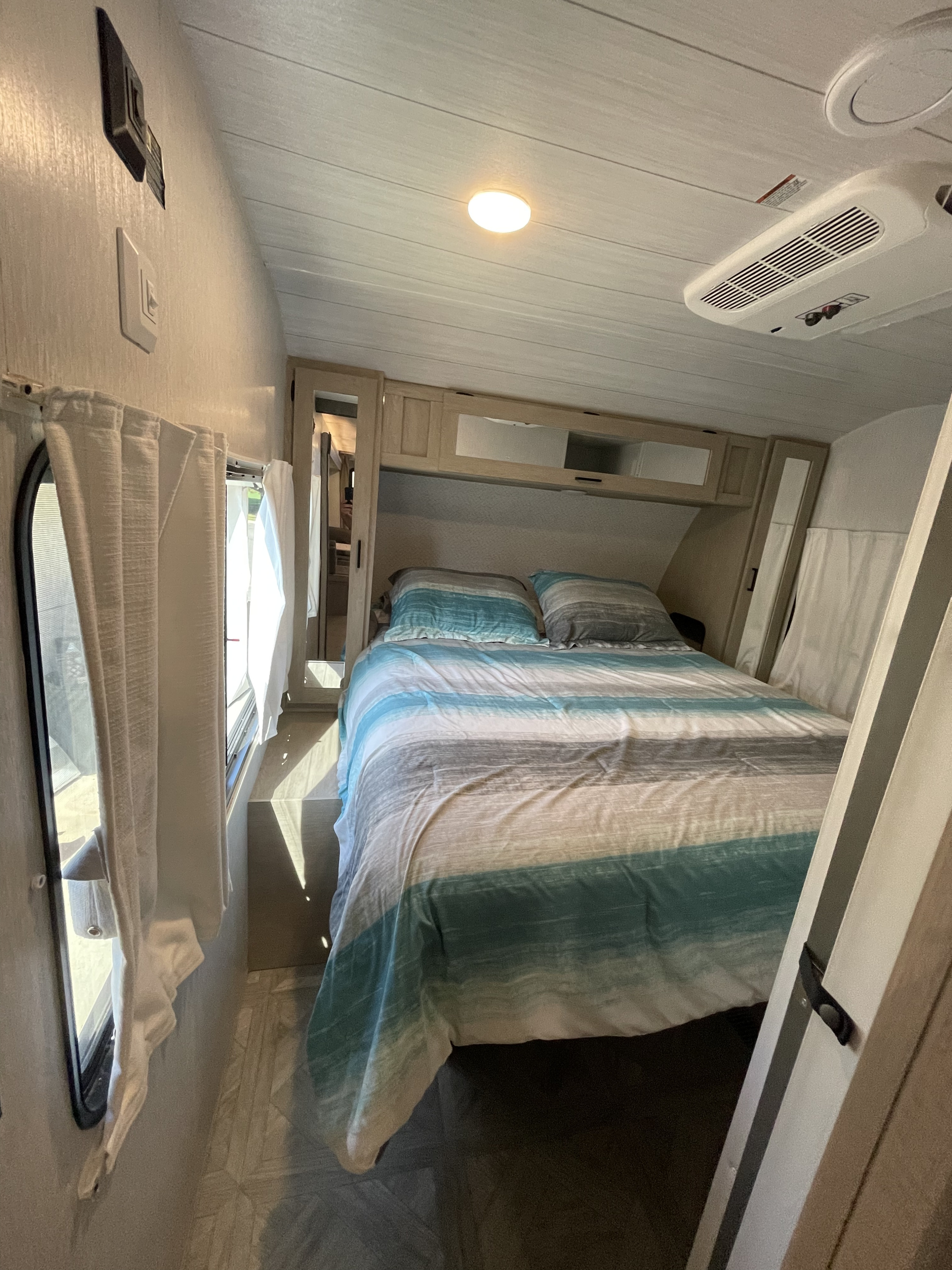 Cozy RV rental bedroom: striped bed, overhead storage, white curtains, and AC, offering comfort on the go.. Other Other 2024