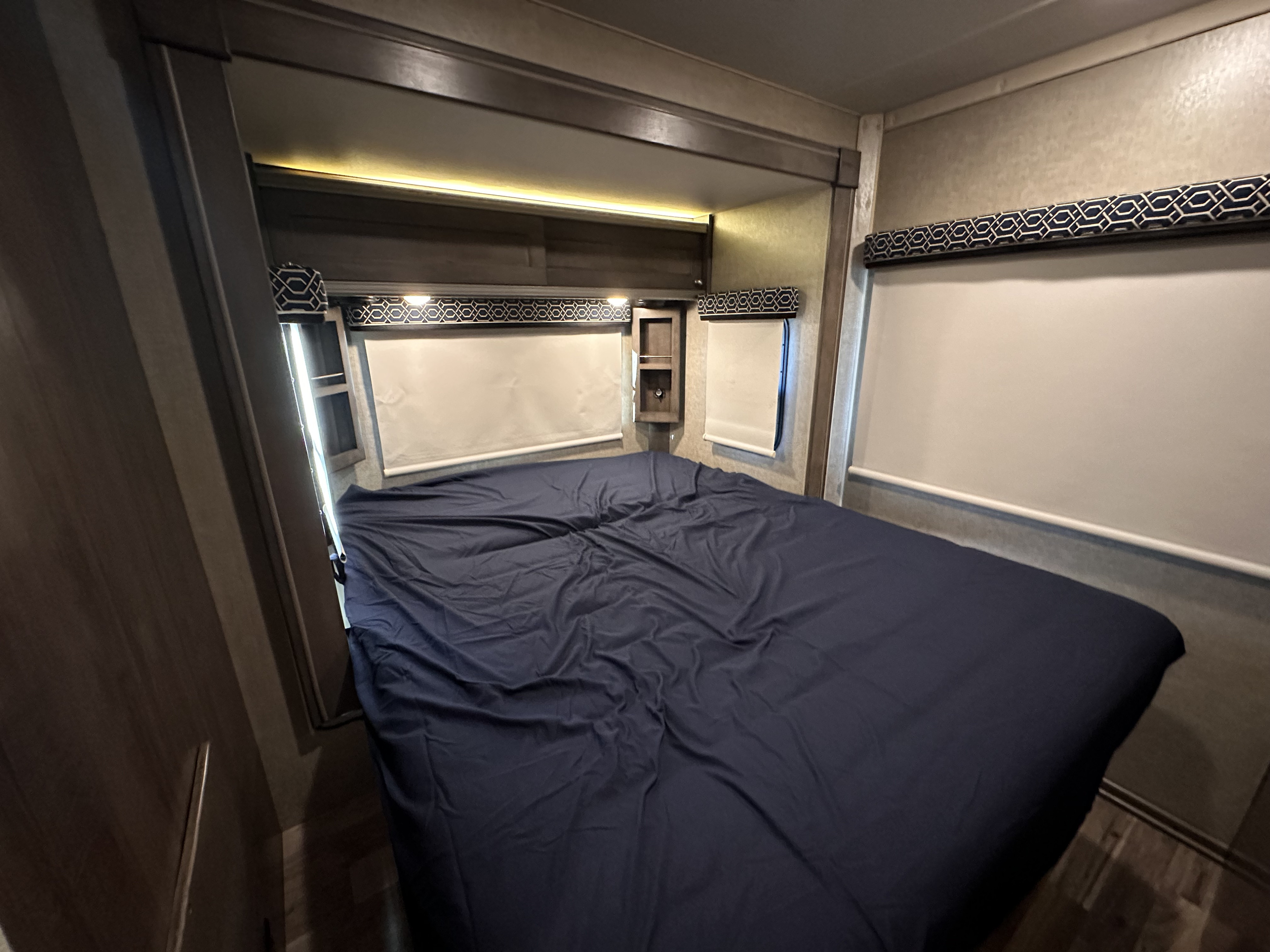 Cozy RV rental bedroom featuring a large bed with dark blue sheets, pull-down blinds, and stylish wooden interior.. Winnebago Fuse 2017