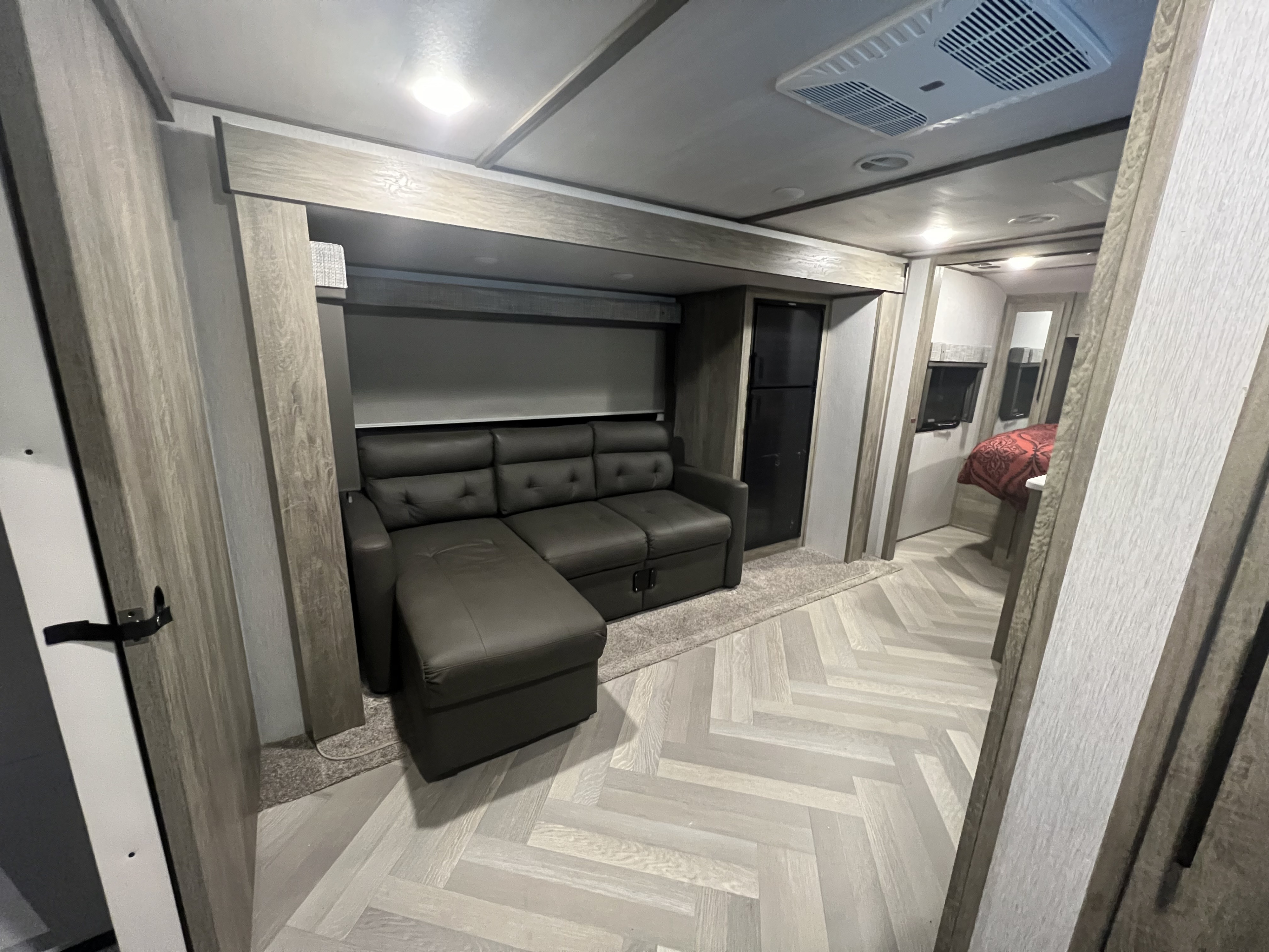 Modern RV rental living area with comfortable sectional sofa and light wood herringbone floor leading to bedroom.. Heritage 22RBHL 2022
