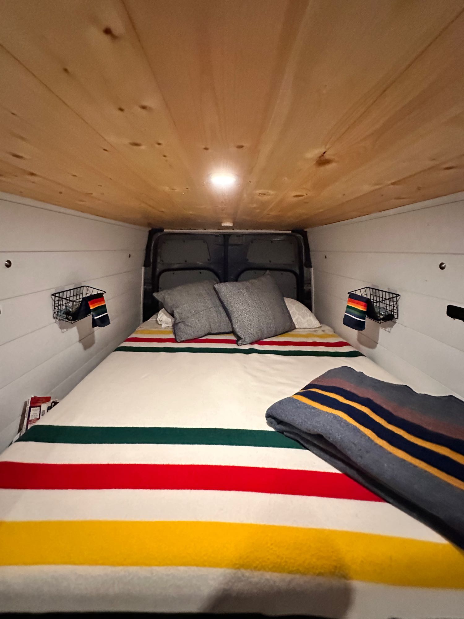 Cozy RV rental camper van interior featuring a comfortable bed with colorful blankets, wood ceiling, and wall baskets.. Mercedes-Benz Sprinter 2020
