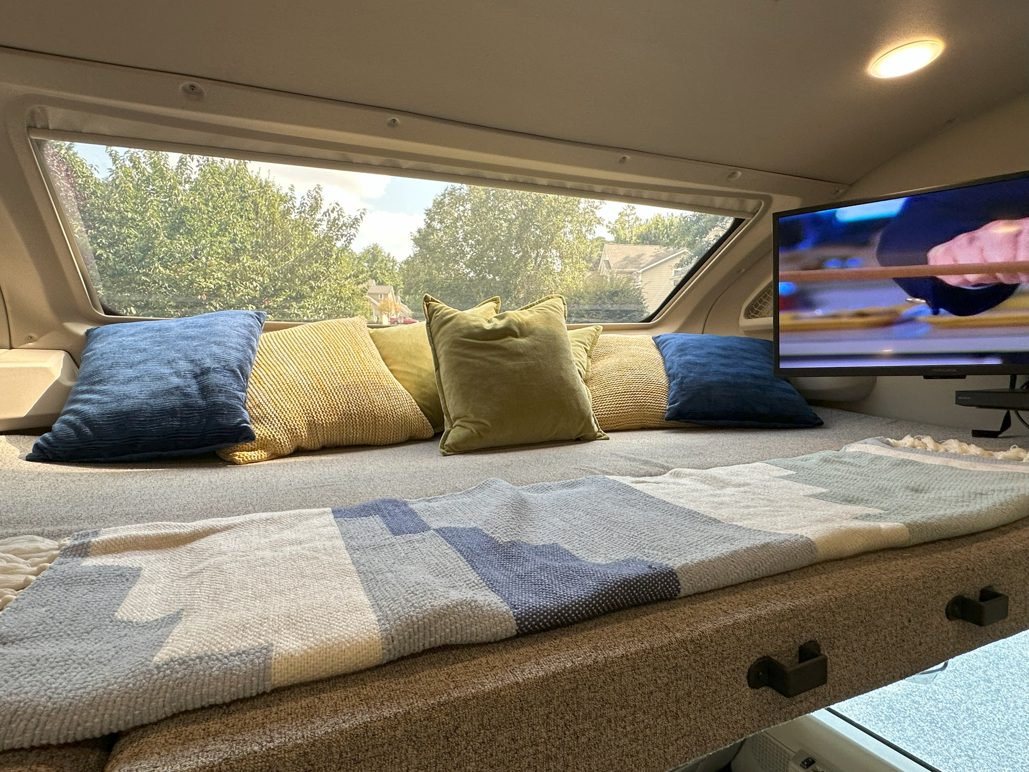Cozy RV rental interior: Bed with colorful pillows, throw blanket, TV, and a large window view of nature.. Entegra Coach Esteem 2021