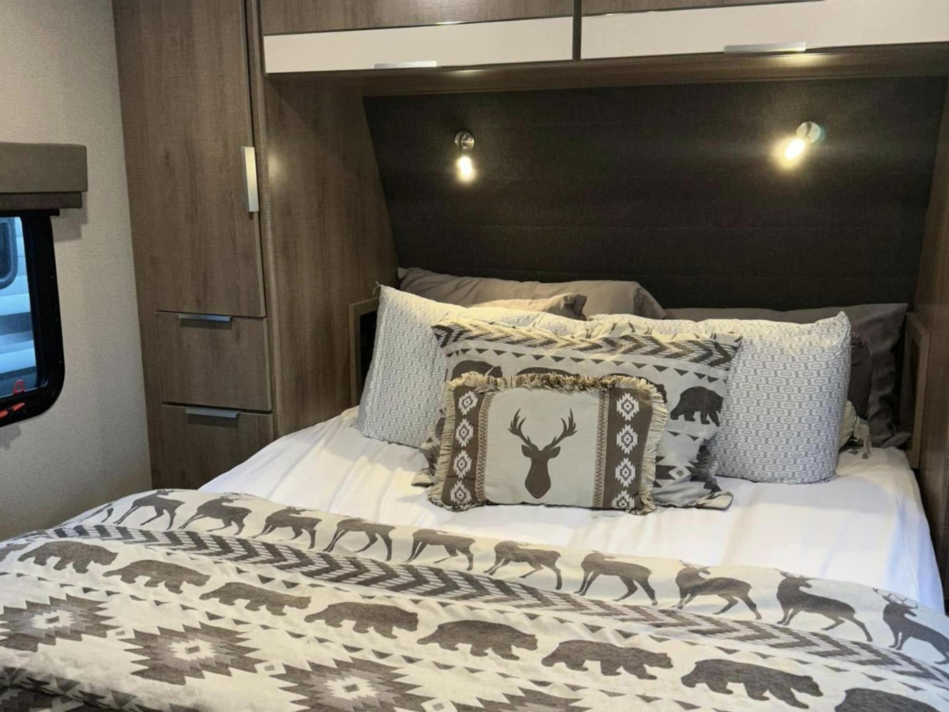 This RV rental bedroom offers a cozy bed with nature-themed bedding, built-in storage, and soft lighting for travel comfort.. Grand Design Imagine XLS 2023