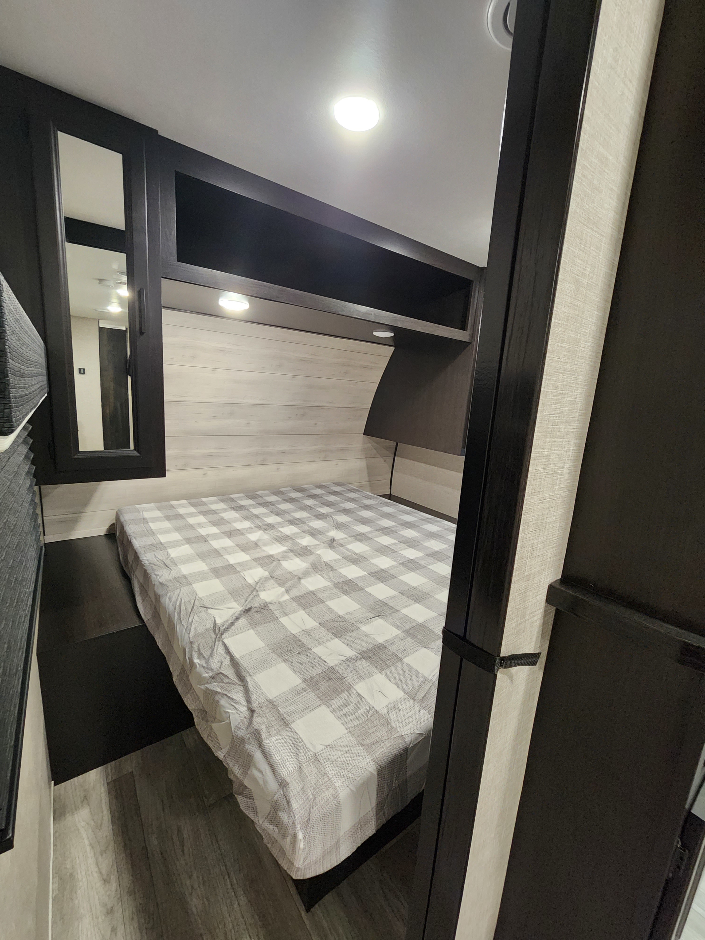 Clean & compact RV rental bedroom. Features a plaid bed, dark wood cabinets, light wood walls, & overhead lighting/storage.. Jayco Jay Flight 2023