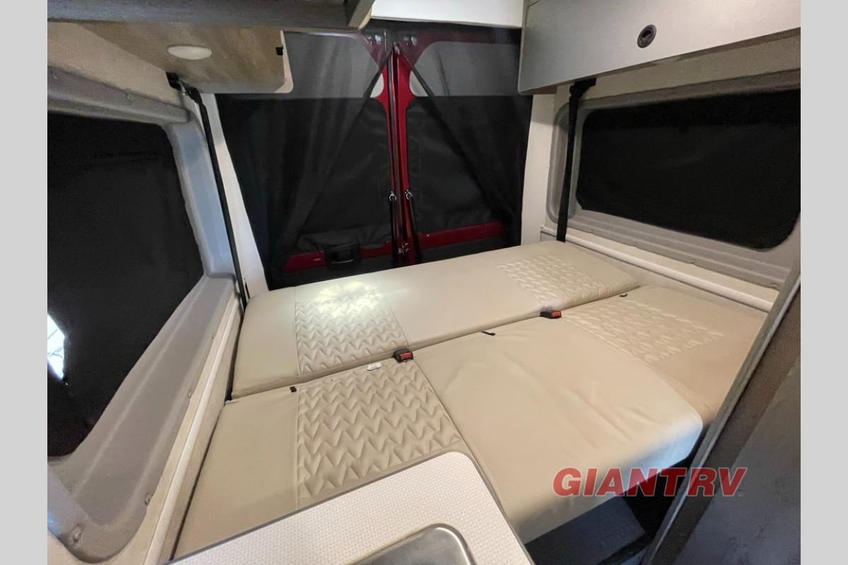 This RV rental interior shows a convertible bed with textured beige cushions, black privacy screens on windows and rear doors, and overhead cabinets.. Winnebago Solis 2022