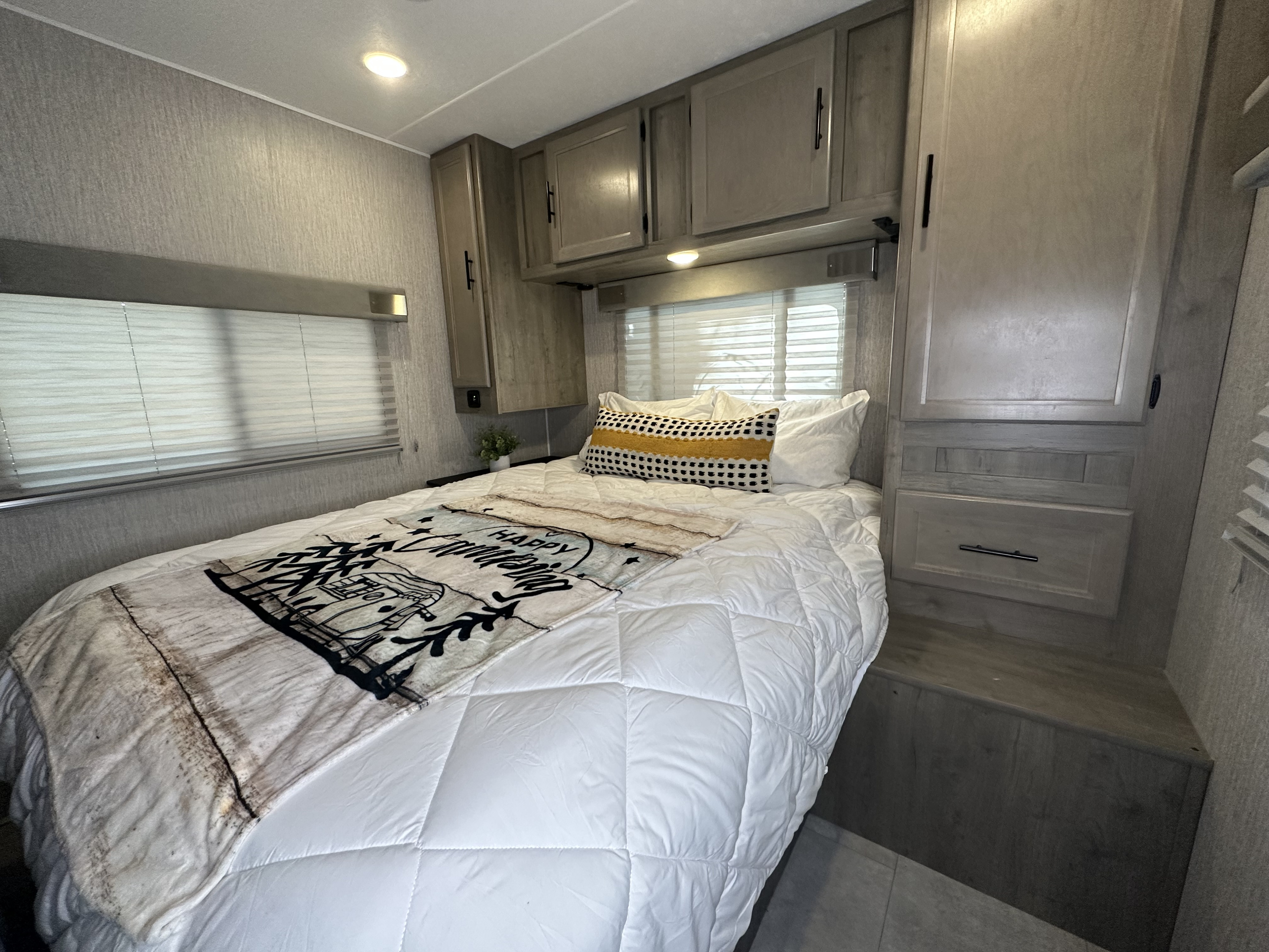 Inside this comfy **RV rental**: a bedroom with a cozy bed, 'Happy Camper' throw, window blinds, and ample storage.. Forest River Coachmen Freelander 2022