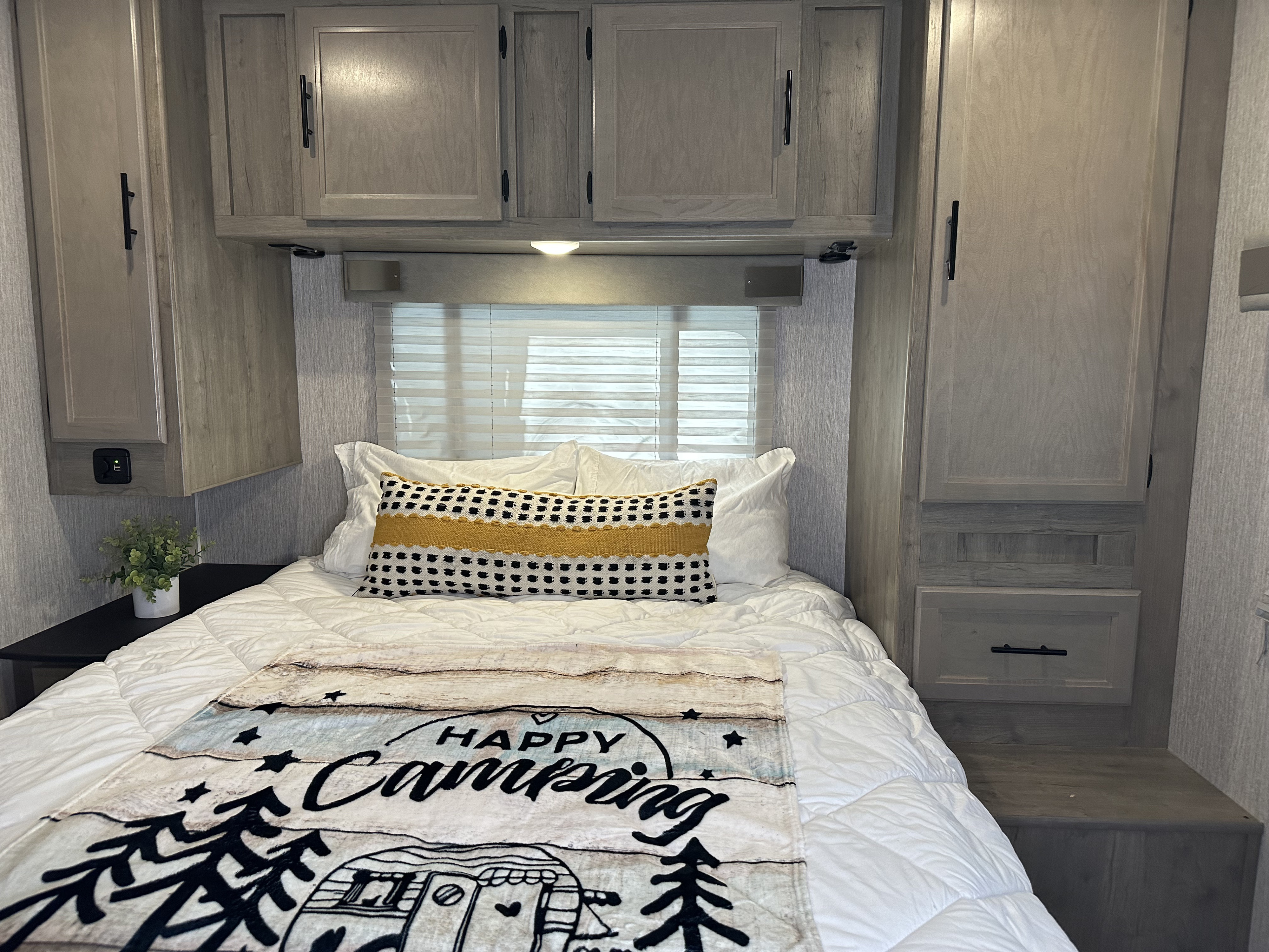 Cozy RV rental bedroom featuring a clean bed, "Happy Camping" blanket, and light wood-look storage cabinets.. Forest River Coachmen Freelander 2022