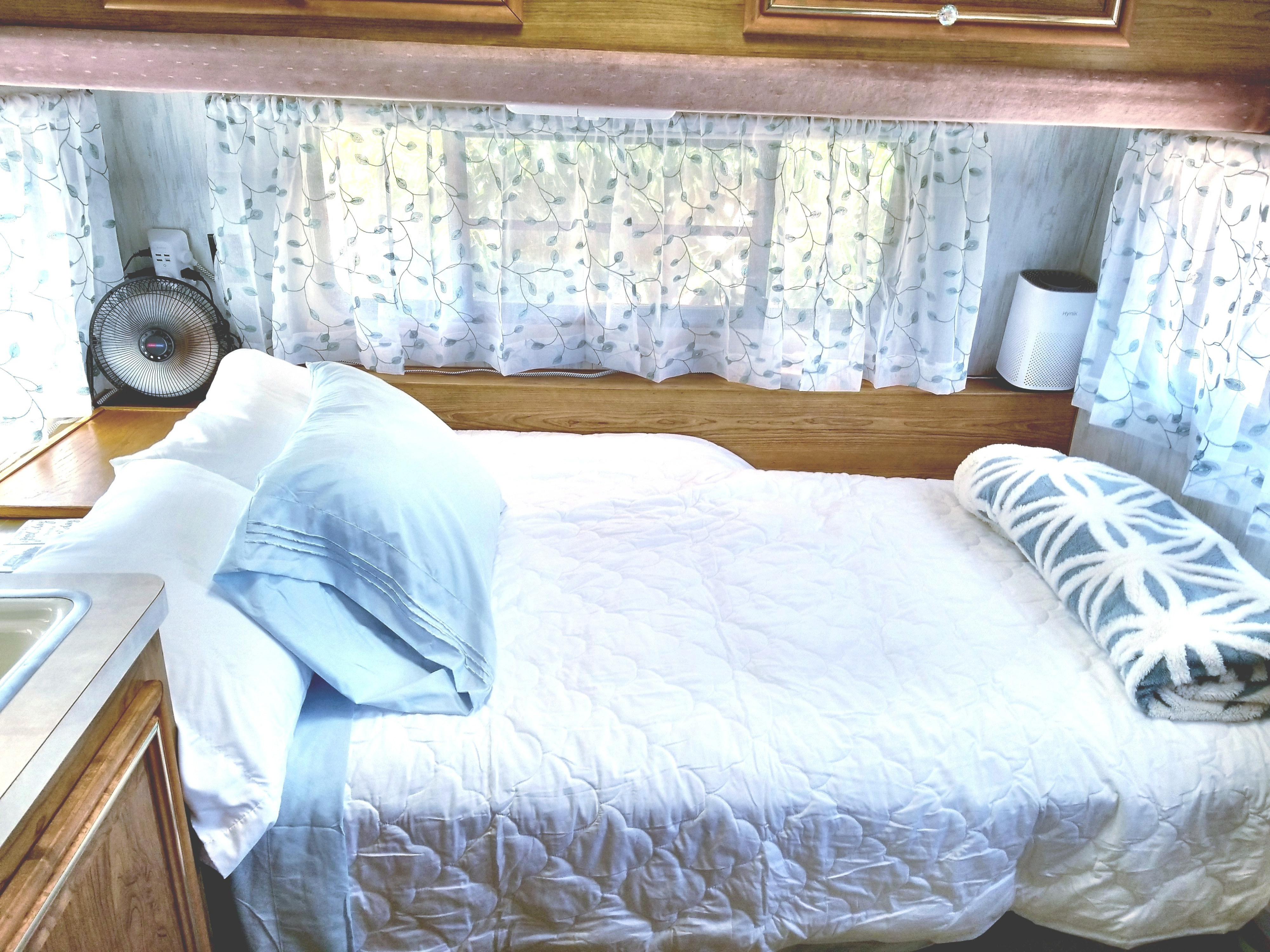 RV rental interior featuring a neatly made bed with white and blue bedding, sheer leaf-patterned curtains, a fan, and an air purifier.. Fleetwood Prowler 1990