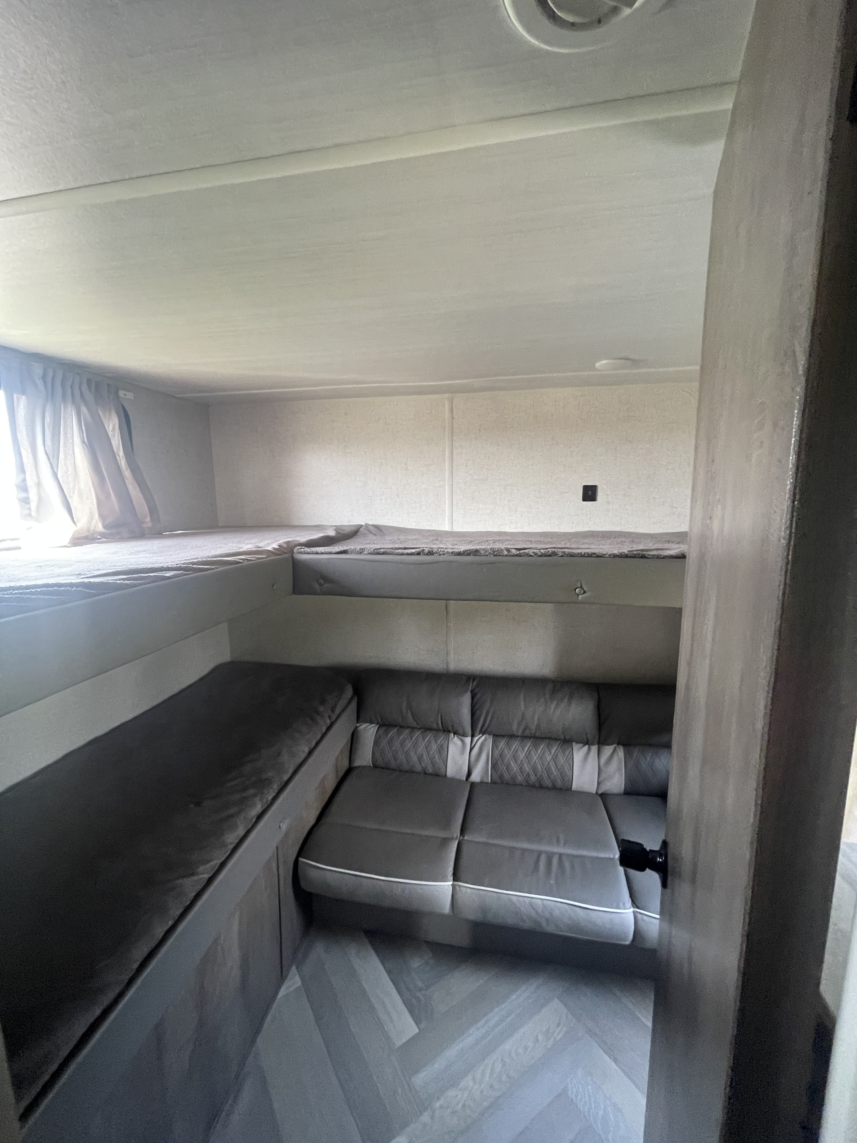 RV rental interior features a cozy lower sofa/dinette, an upper bunk bed, neutral curtains, and herringbone flooring.. Forest River Wildwood X-Lite 2021