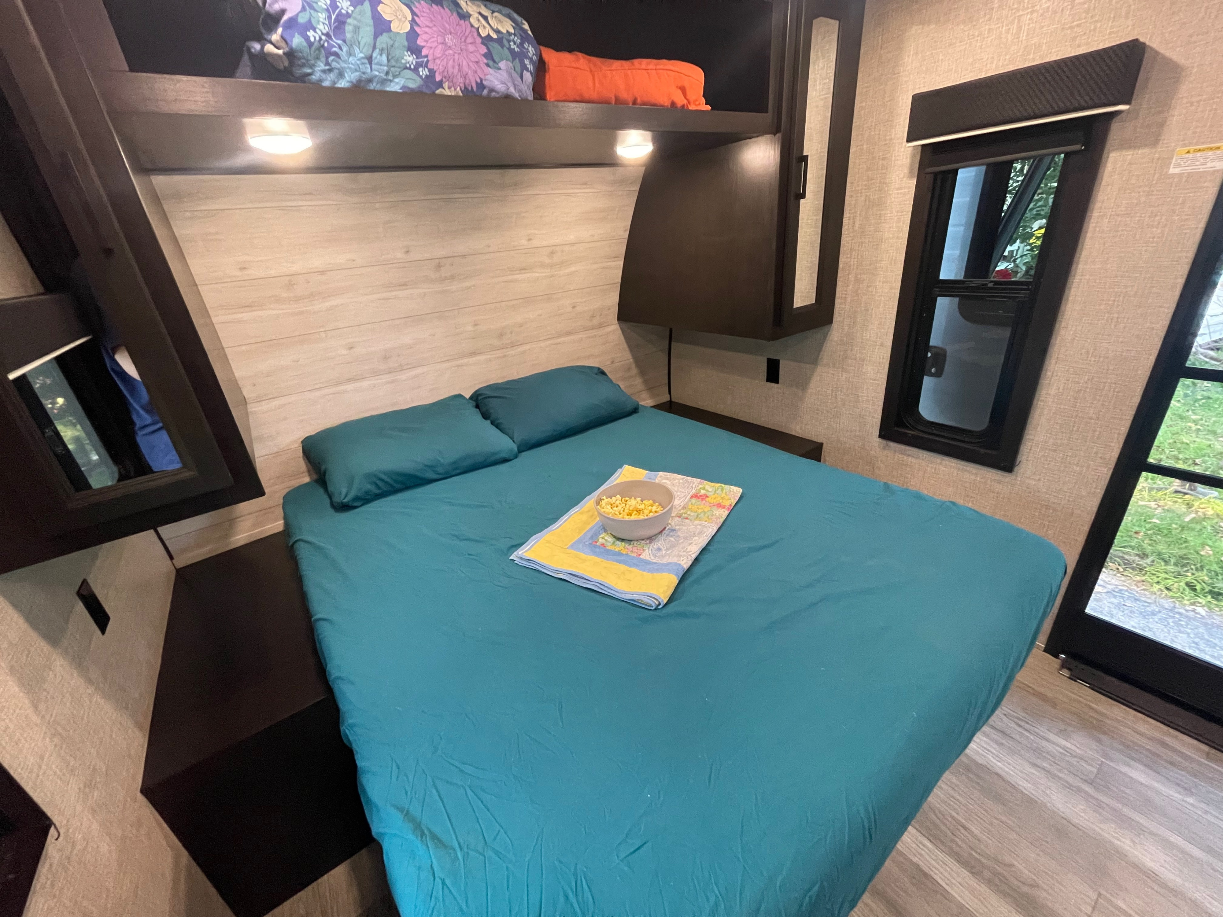 Comfortable RV rental bedroom with teal bed, pillows, and popcorn bowl. Features overhead storage, window, and wood accents.. Jayco Jay Flight 2023