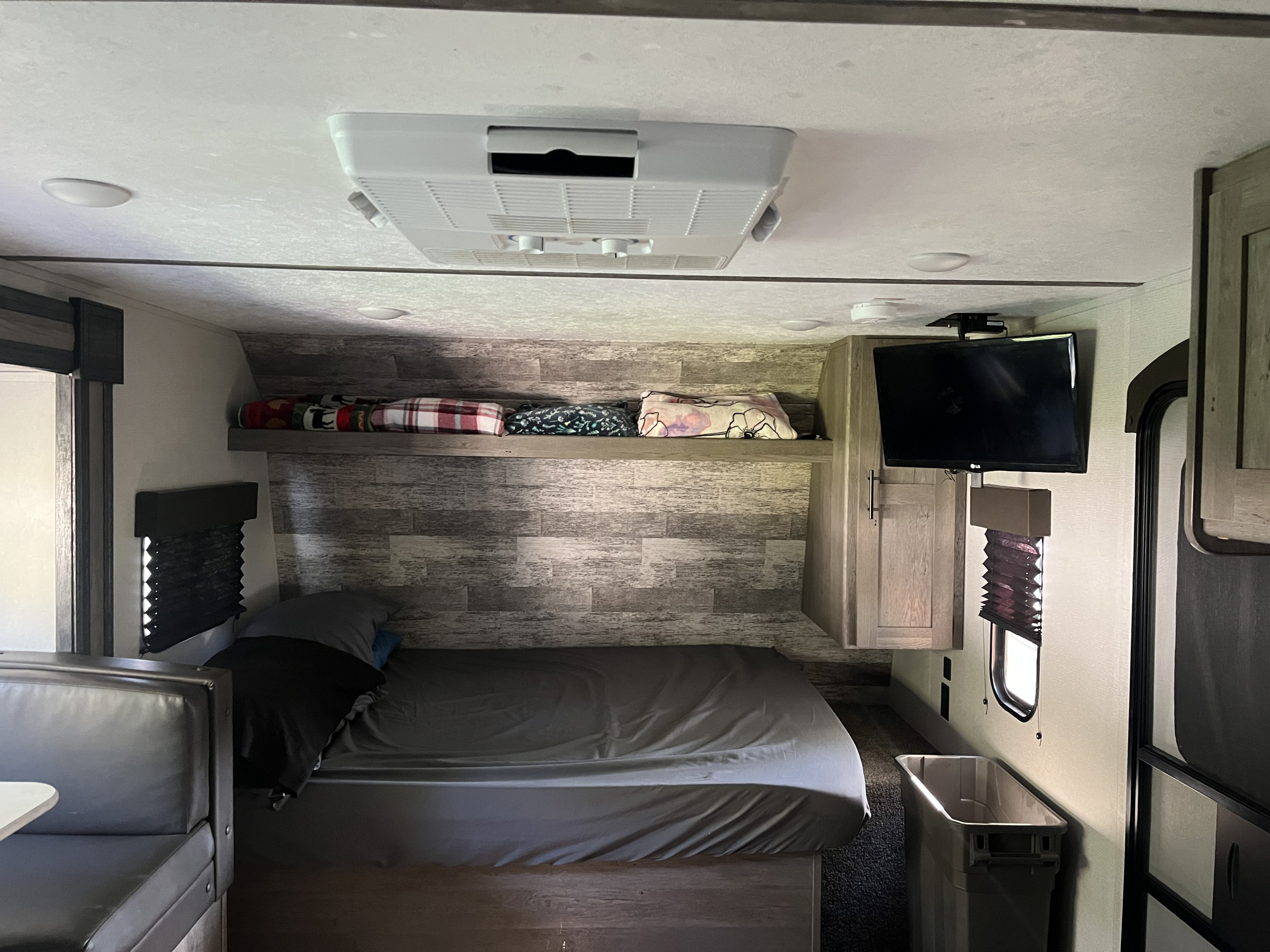 This RV rental interior features a cozy bed with wood-paneled accent wall, dinette, TV, and ceiling AC.. Keystone RV Springdale 2022