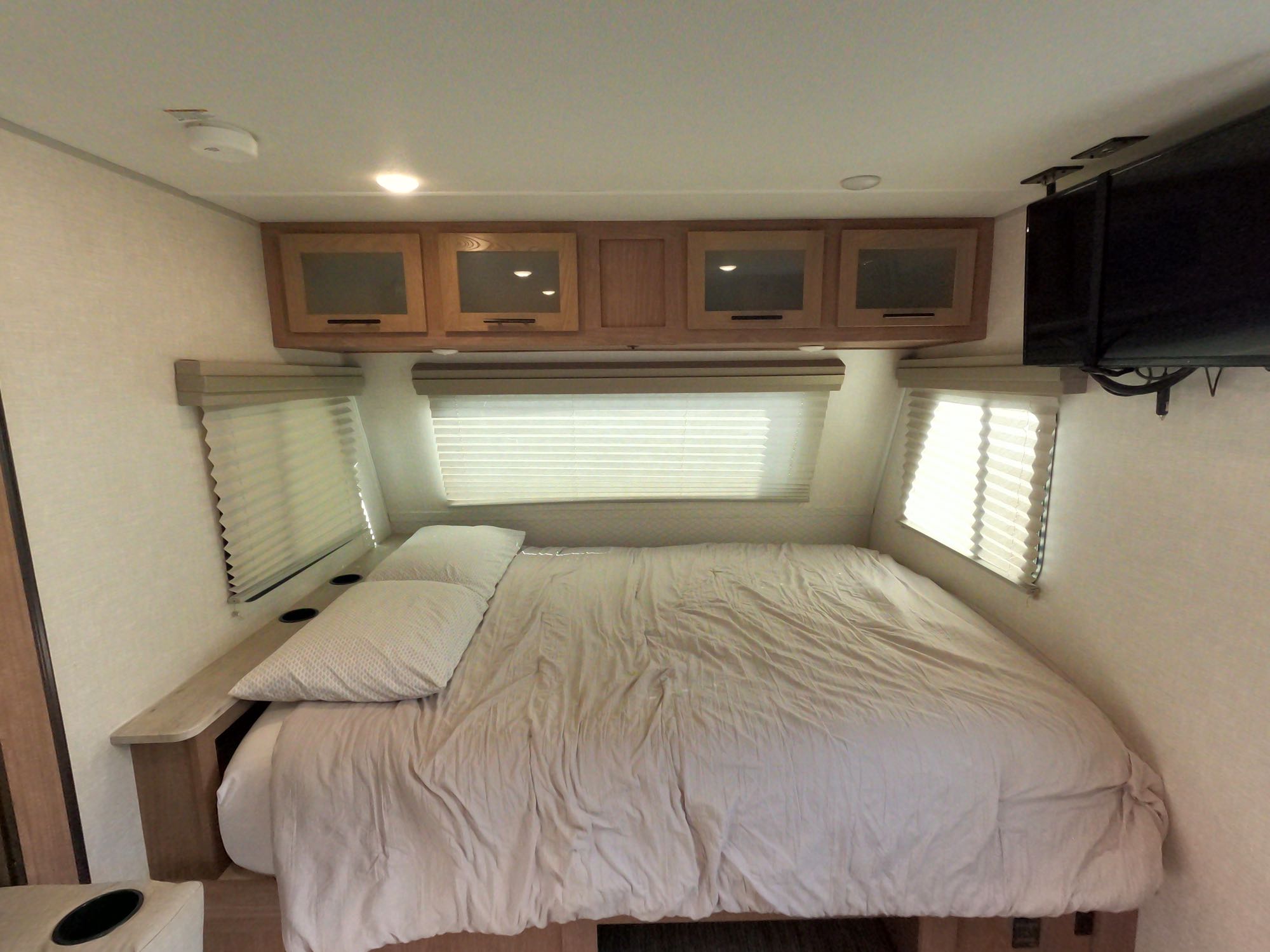 Cozy RV rental bedroom featuring a large bed, overhead cabinets, window blinds, and a ceiling-mounted TV.. No Boundaries 19.8 2021