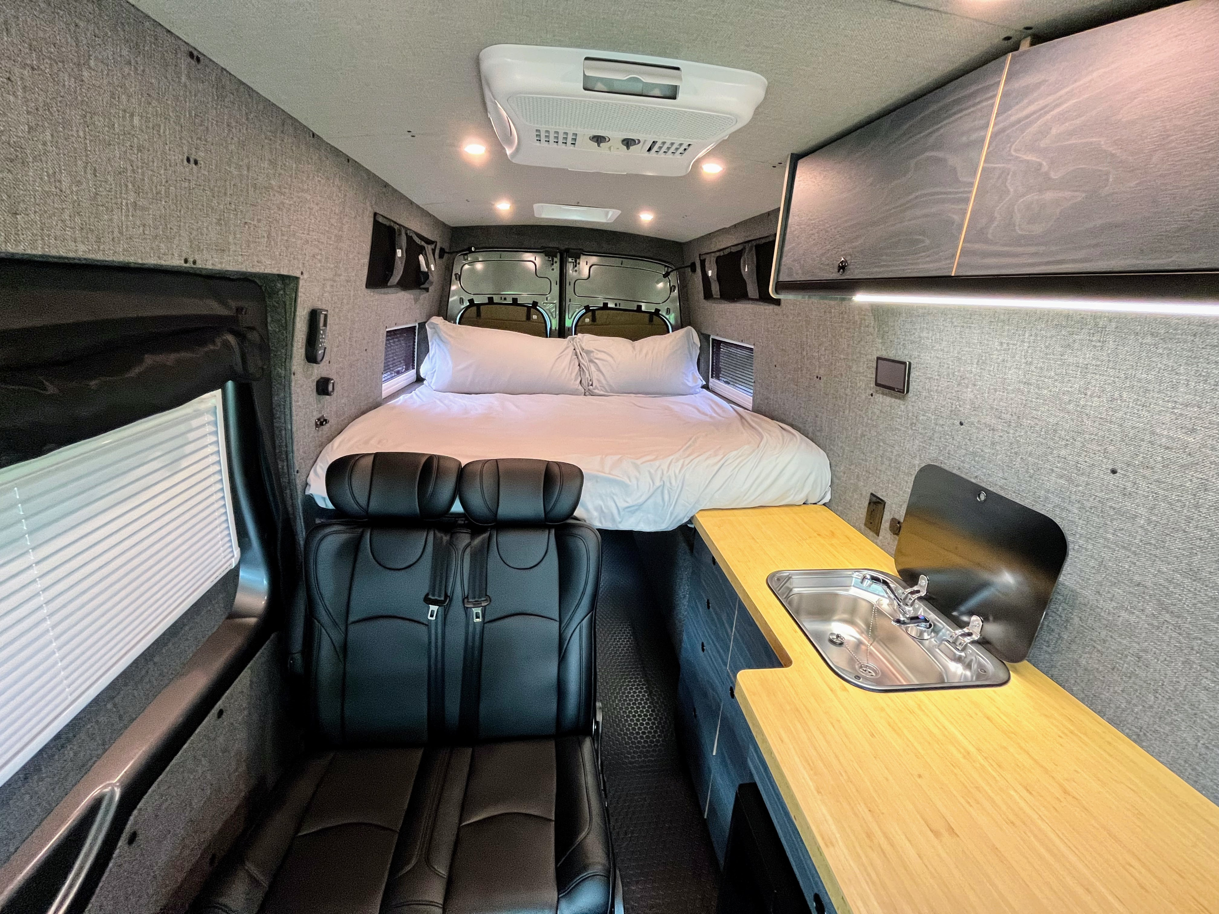 Interior of a modern RV rental camper van, featuring a bed at the rear, a kitchen counter with sink, and passenger seating.. Ford Custom Transit 2022
