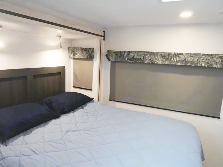 RV rental bedroom with light blue bed, dark headboard, and windows featuring gray roller shades and patterned valances.. Forest River Sunseeker Classic 2023