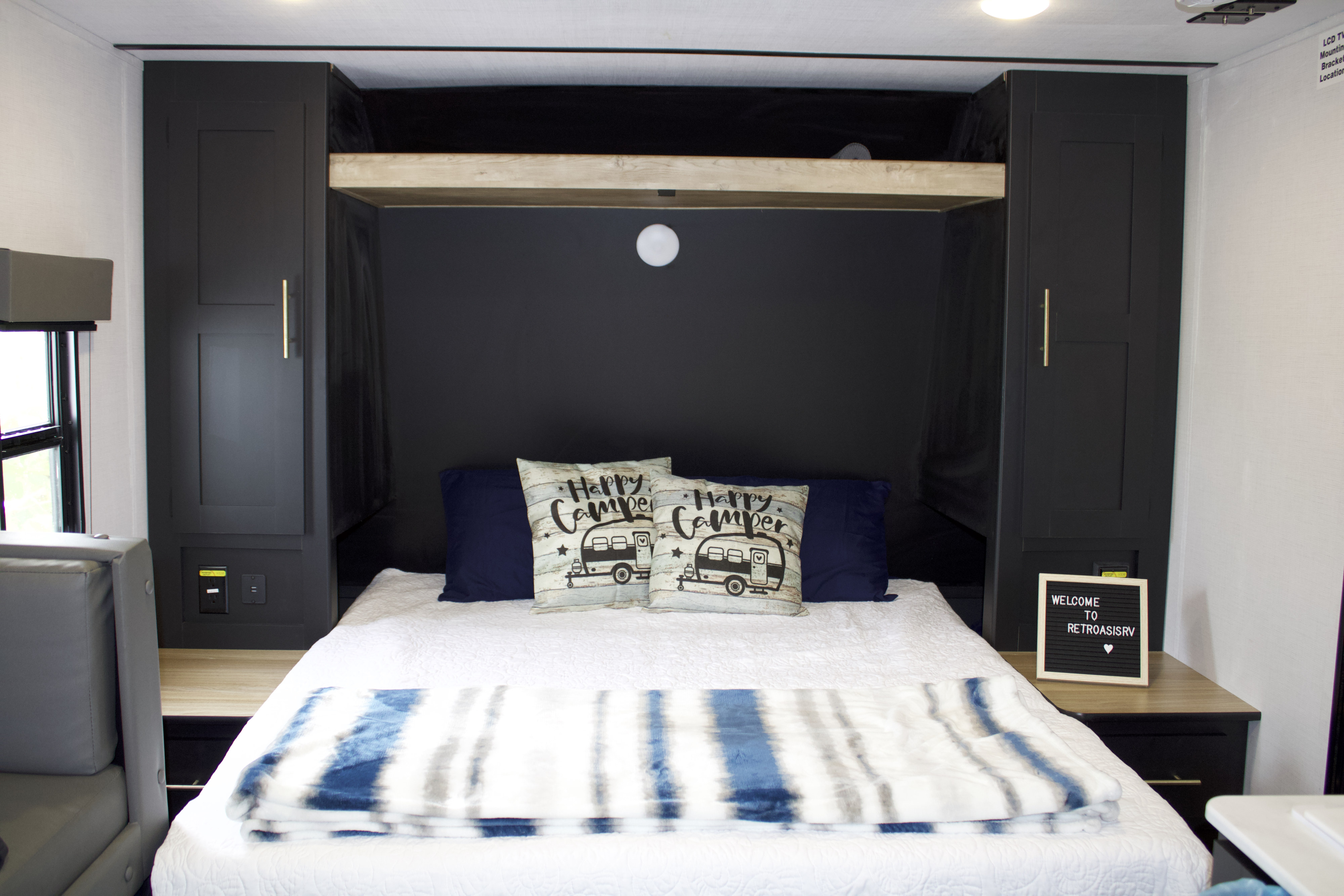A cozy RV rental bedroom featuring a white bed with "Happy Camper" pillows, dark storage cabinets, and a welcoming ambiance.. Keystone RV Springdale 2023