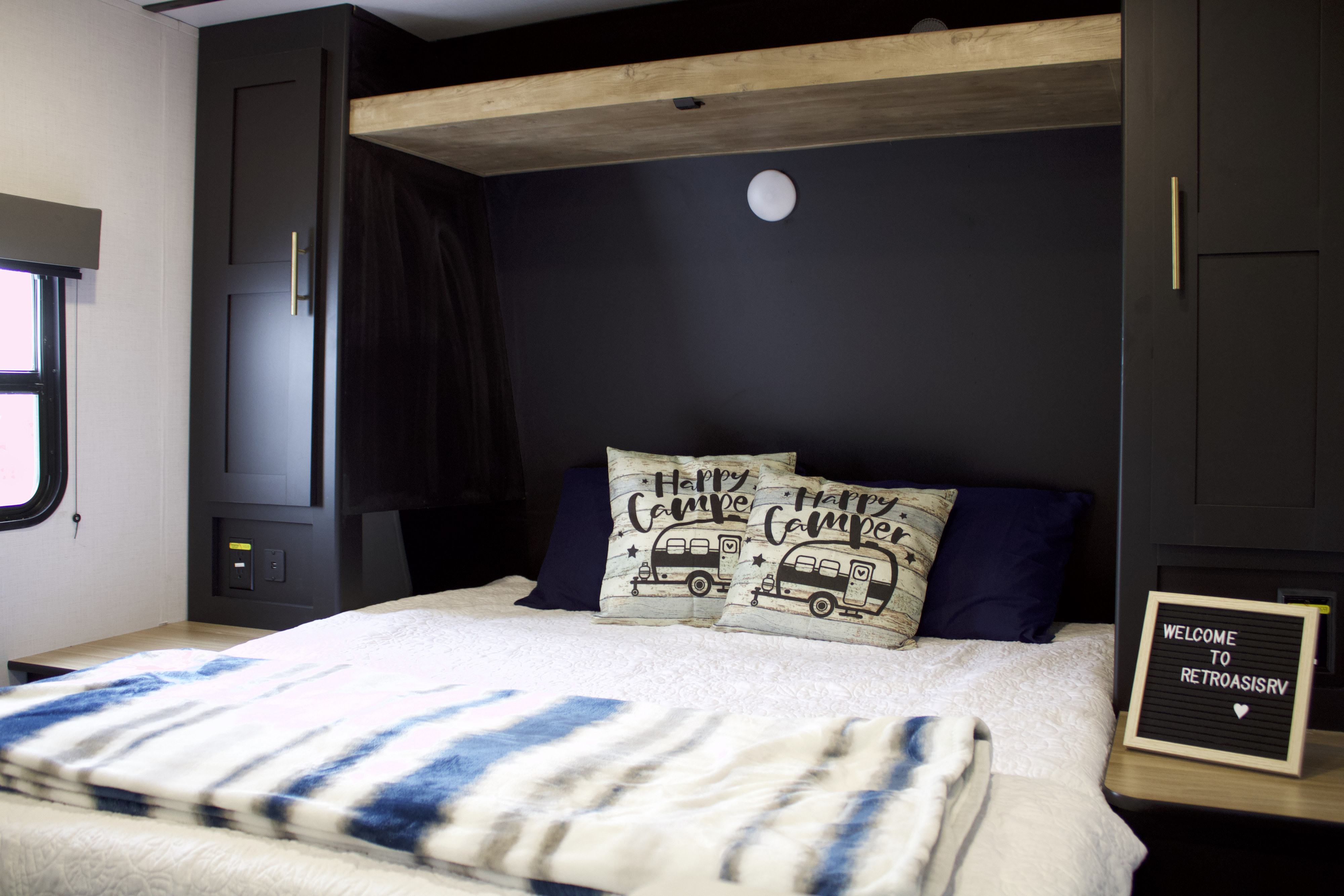 Modern RV rental bedroom featuring a comfy bed, "Happy Camper" pillows, black cabinetry, and a friendly welcome sign.. Keystone RV Springdale 2023