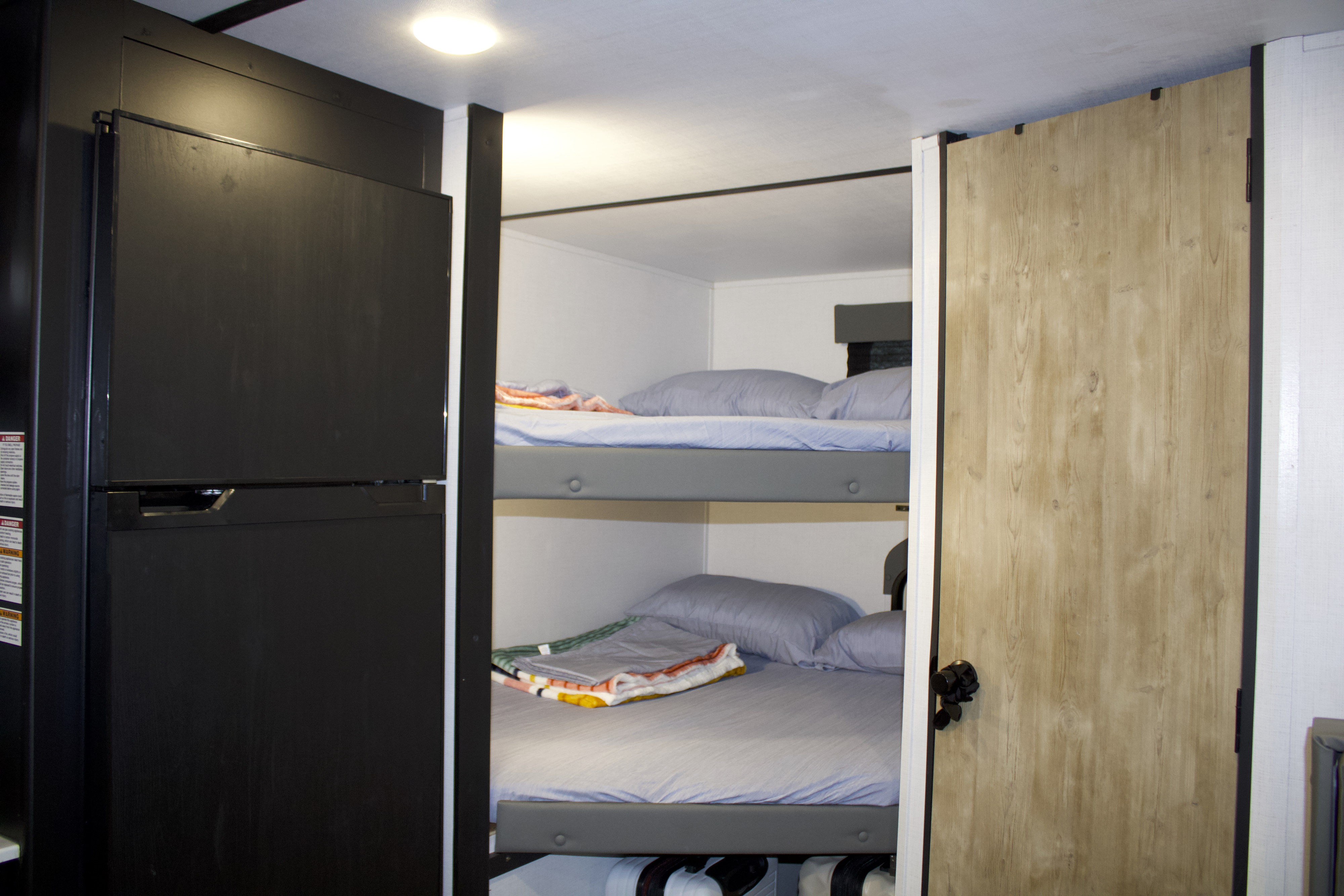 Inside an RV rental: dark refrigerator, stacked bunk beds with blue bedding, and a light wood door.. Keystone RV Springdale 2023