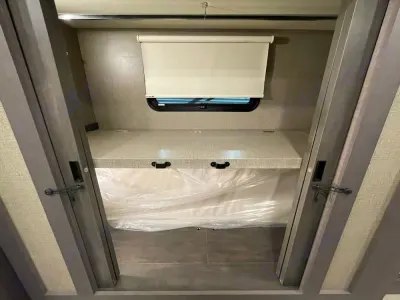 RV rental interior: A compact sleeping bunk with window, blind, shelf, and new mattress in plastic.. Thor Miramar 2022