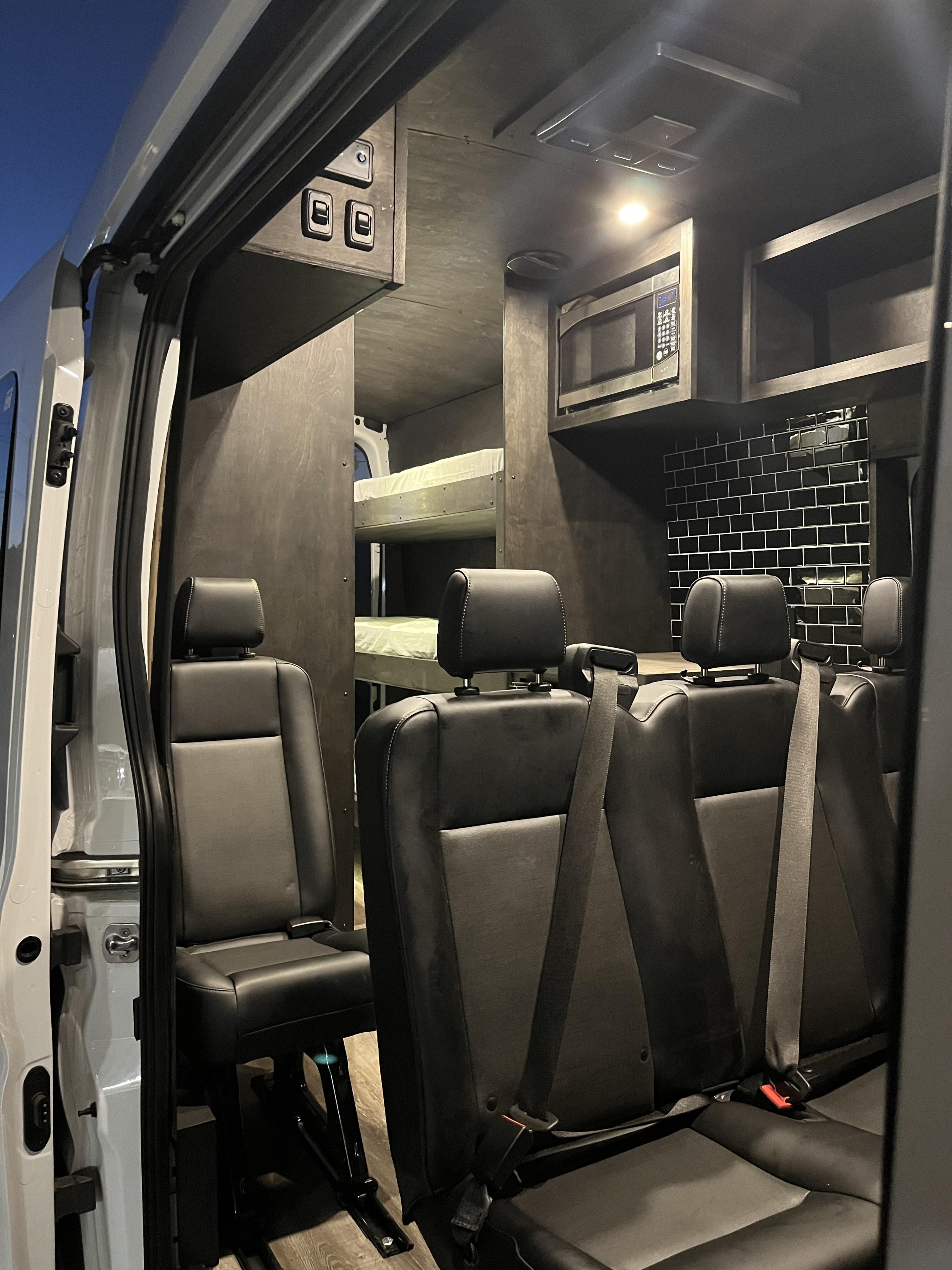 View inside a modern rv rental with black passenger seats, bunk beds, and a compact kitchen setup.. FORD 350 HR TRANSIT 2023