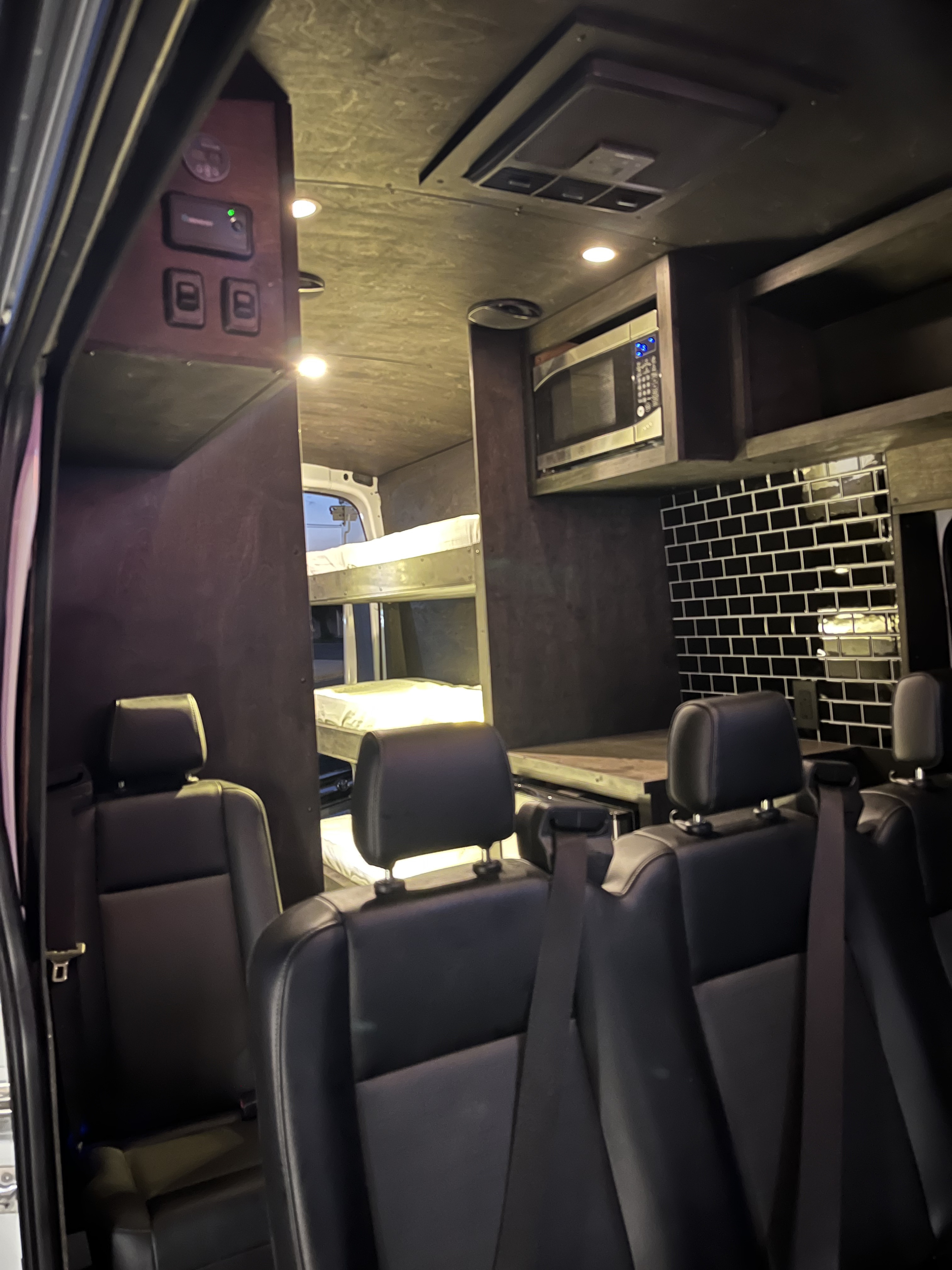 Spacious RV rental interior: bunk beds, passenger seating, and compact kitchen with a microwave.. FORD 350 HR TRANSIT 2023