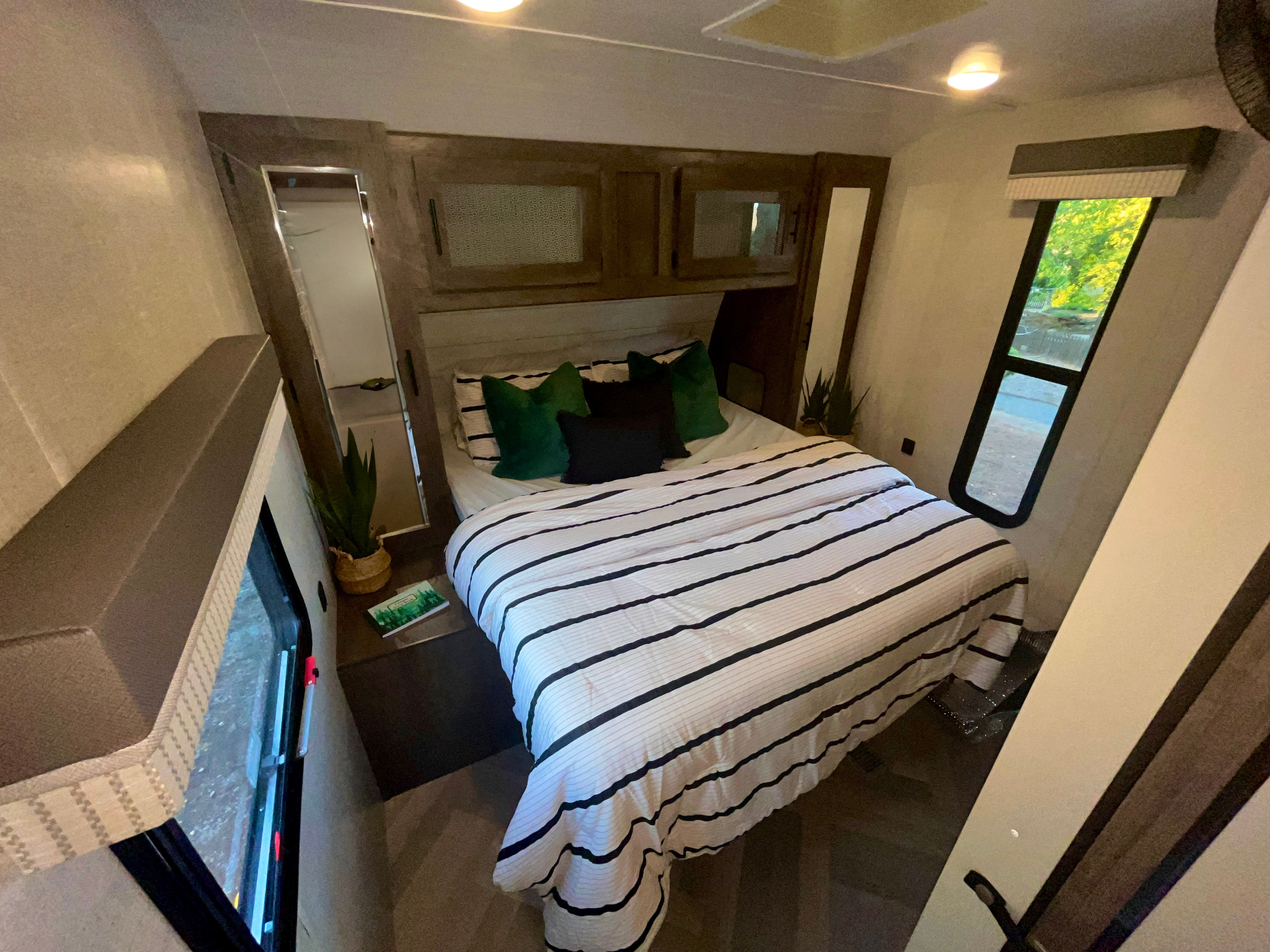 Cozy RV rental bedroom featuring striped bedding, overhead cabinets, and a window with a green outdoor view.. Forest River Wildwood 2021